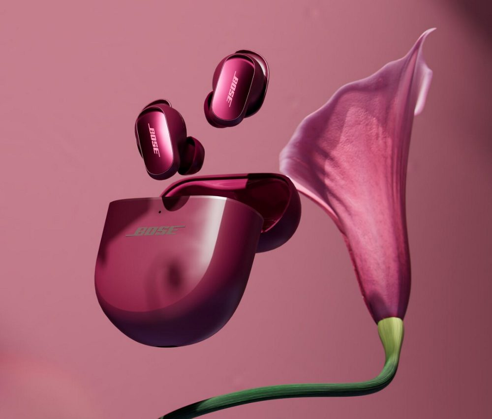 Różowe słuchawki i etui Bose, z różowym kwiatem na różowym tle. Słuchawki unoszą się. Etui jest otwarte.