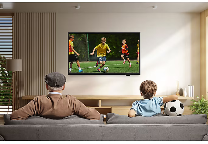 Egy idős férfi és egy gyerek focimeccset néz a tévében. Egy focilabda van a gyerek mellett. A szobában világos fa burkolat van.