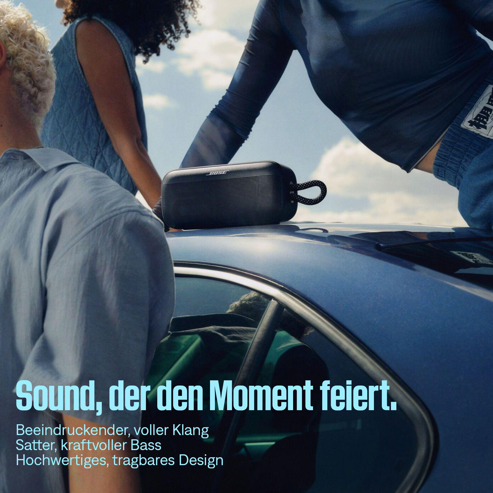 Ein schwarzer Bose-Lautsprecher liegt auf einem blauen Auto. Drei Personen sind zu sehen. Der Himmel ist sichtbar.