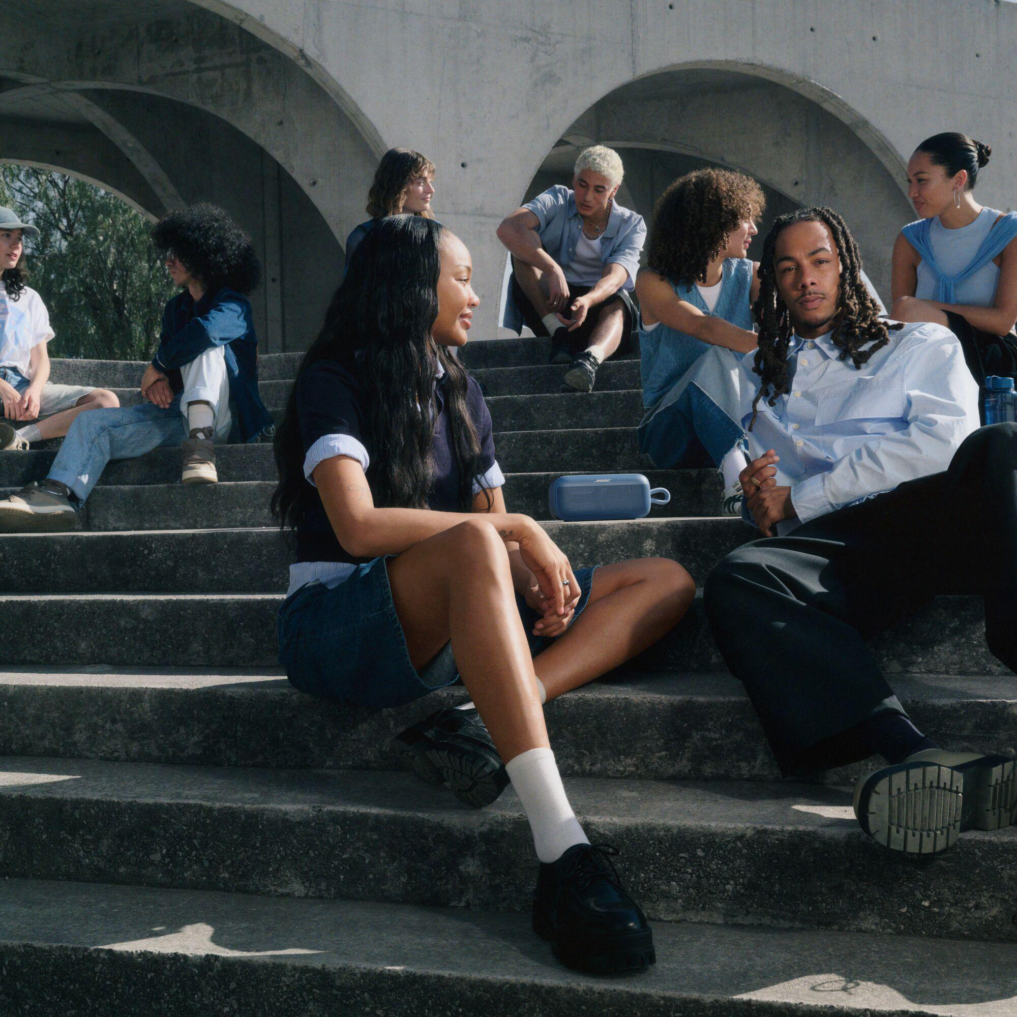 Eine Gruppe junger Leute sitzt auf Betontreppen und unterhält sich. Einige tragen Jeans. Zwischen zwei Personen steht ein Lautsprecher.