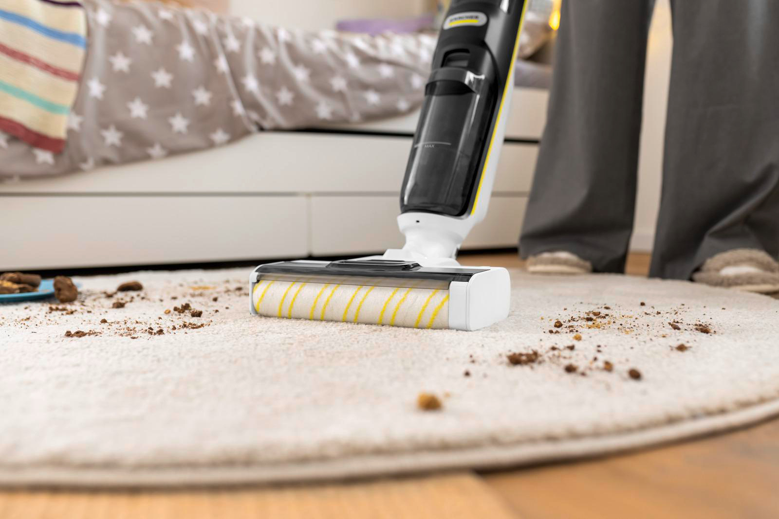 Eine Person saugt einen schmutzigen Teppich mit einem kabellosen Staubsauger. Der Teppich hat Essensreste. Ein Bett mit einer sternförmigen Bettdecke ist sichtbar.