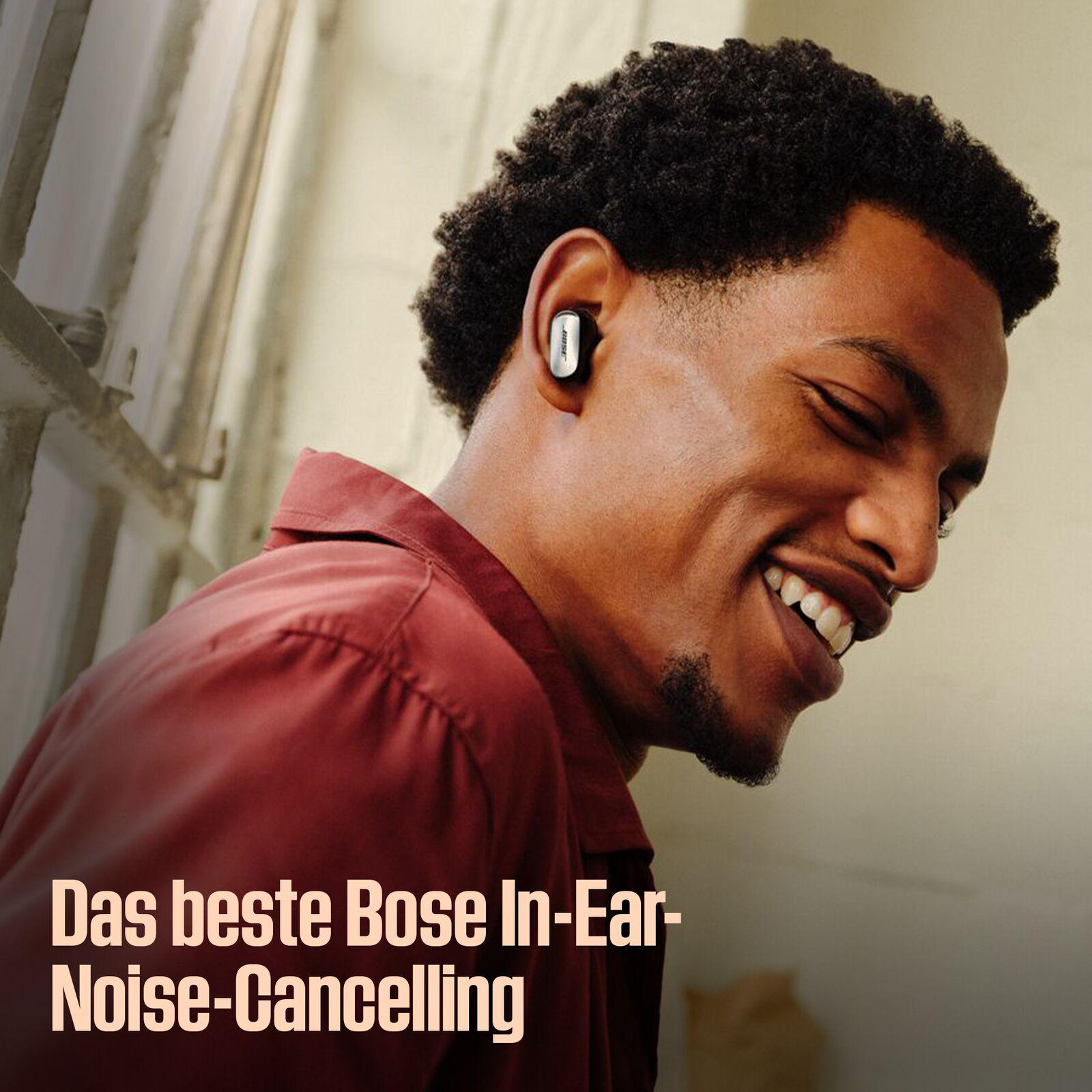Lächelnder schwarzer Mann mit rotem Hemd, Bose-Ohrhörer sichtbar. Text: Das beste Bose In-Ear-Noise-Cancelling.