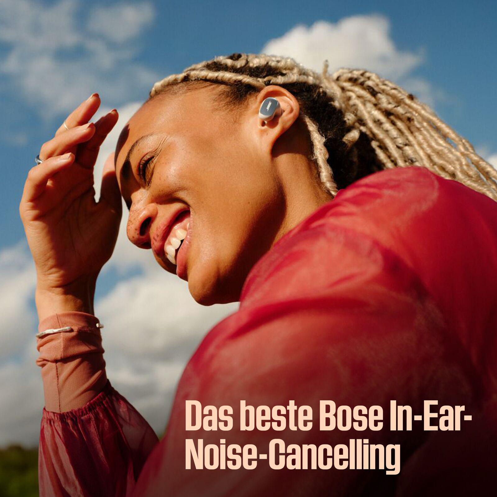 Eine lächelnde Frau mit blonden Dreadlocks trägt ein rotes Oberteil und einen blauen Ohrhörer. Blauer Himmel und Text 'Das beste Bose In-Ear-Noise-Cancelling'.