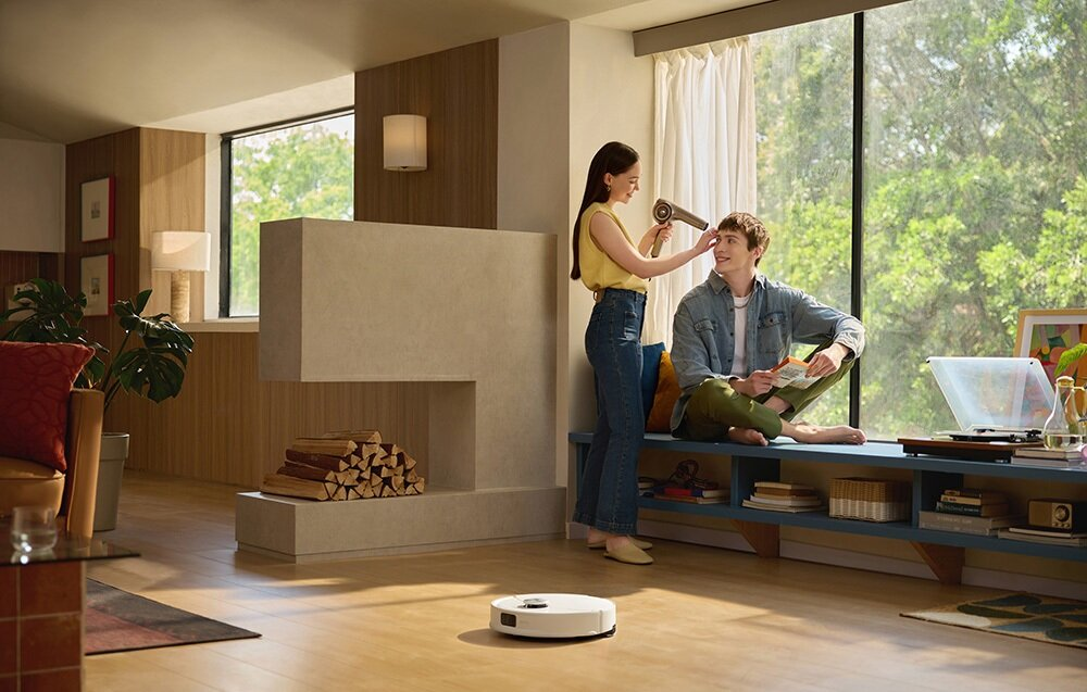Kobieta suszy włosy mężczyzny suszarką. Robot sprzątający czyści podłogę. W tle okno i kominek.