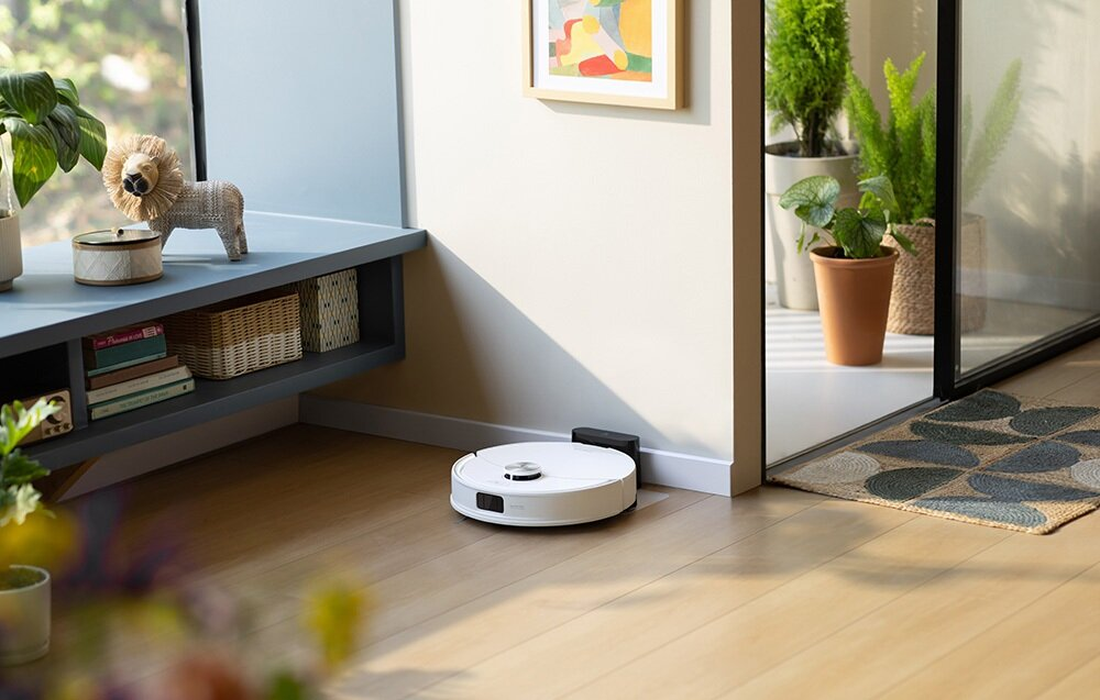 Biały odkurzacz robot na drewnianej podłodze. W pokoju znajduje się również posąg lwa, rośliny i obraz.