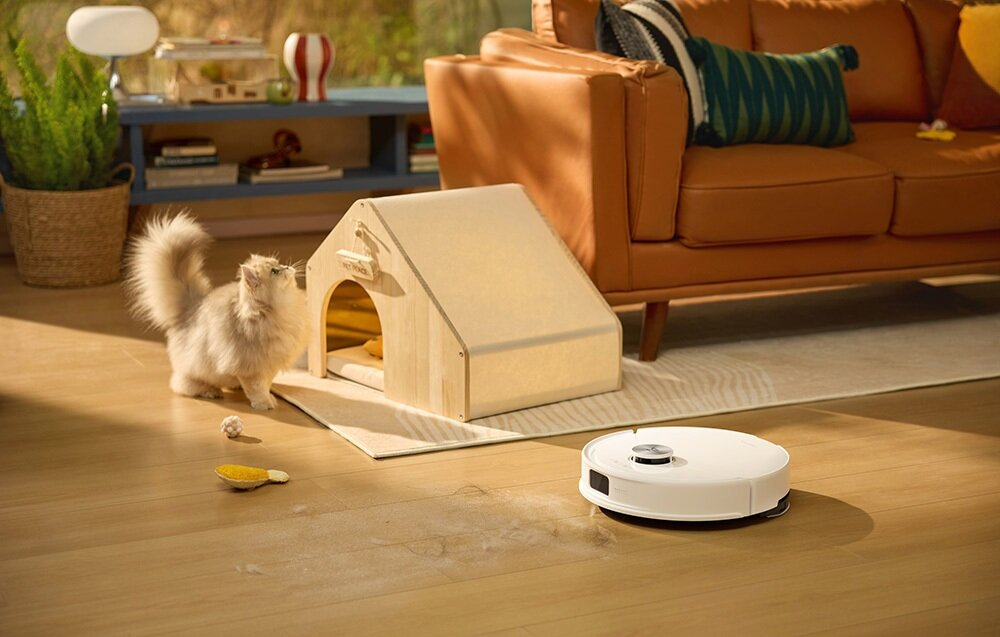 Puszysty kot patrzy na domek dla kota. Odkurzacz robot stoi na podłodze. Kanapa w tle.