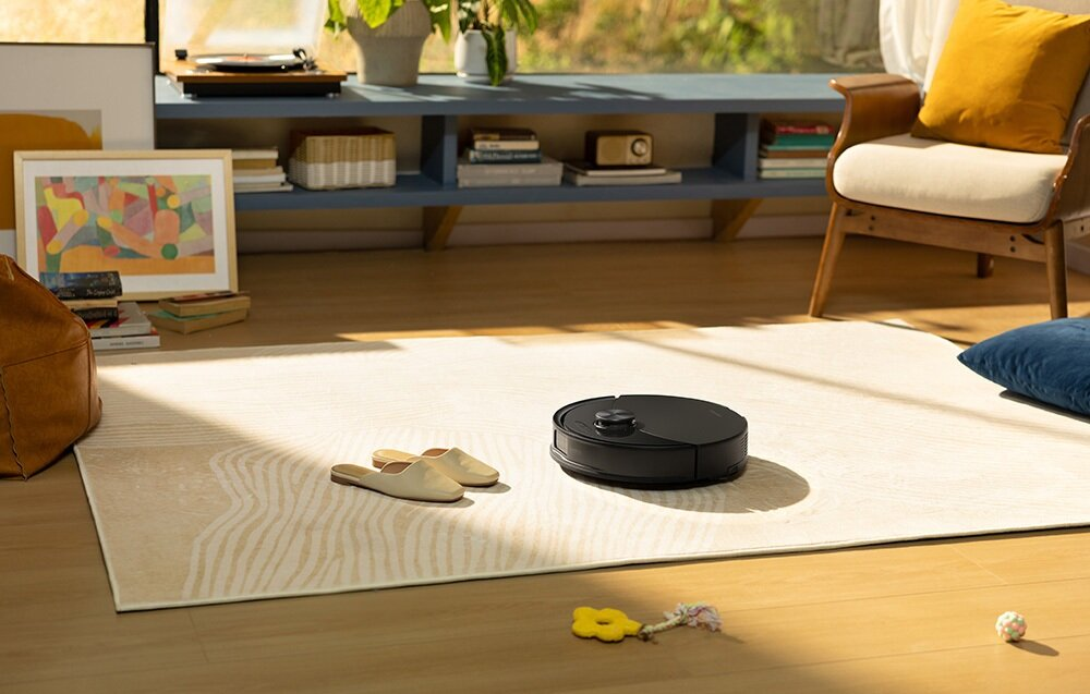 Czarny robot odkurzający na dywanie. Para butów, żółta zabawka i piłka na podłodze. Krzesło i półka w tle.