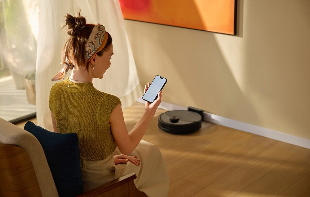 Kobieta trzyma telefon, patrząc na robota sprzątającego. Siedzi blisko okna, z obrazem na ścianie.