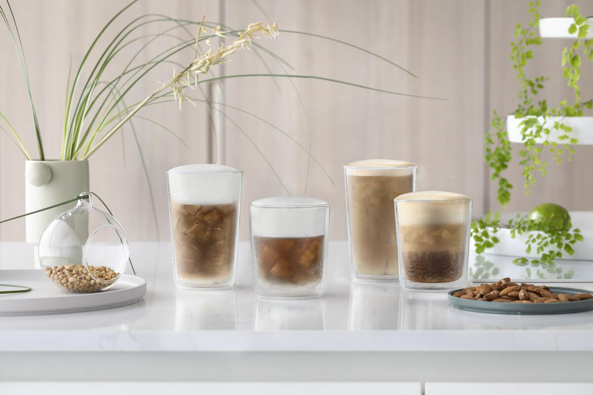 Vier Eiskaffeegetränke mit Schaum, eine Vase mit Pflanzen, eine Glaskugel mit Samen und ein Teller mit Mandeln auf weißer Oberfläche.