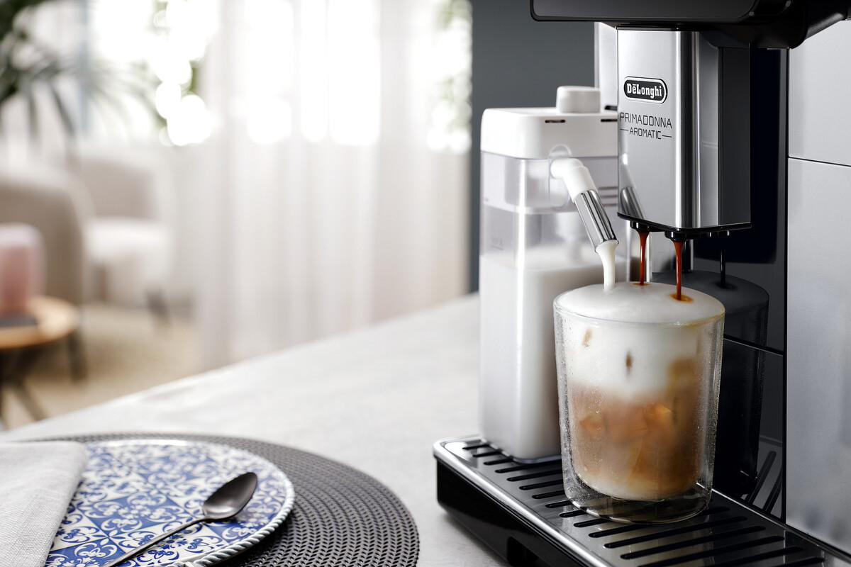 Eine Kaffeemaschine gibt Kaffee und Milchschaum in ein Glas ab. Auf dem Tisch steht ein Teller mit einem Löffel.