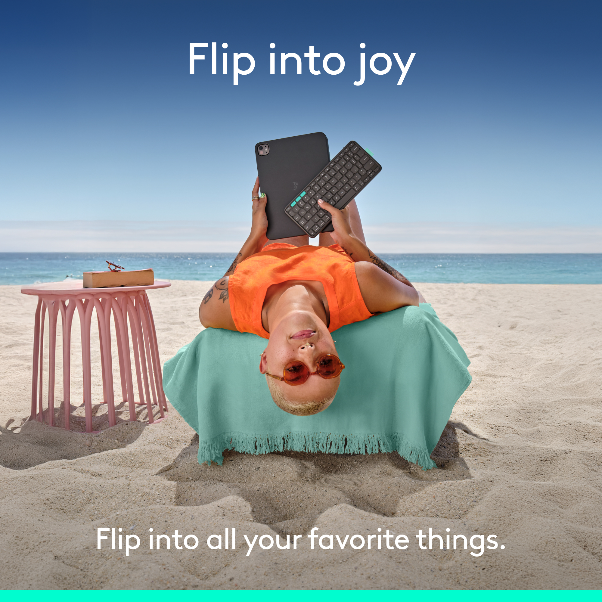 Person am Strand hält Tablet und Tastatur, trägt ein orangefarbenes Oberteil und Sonnenbrille. Rosa Tisch in der Nähe. Blauer Himmel und Ozean im Hintergrund.