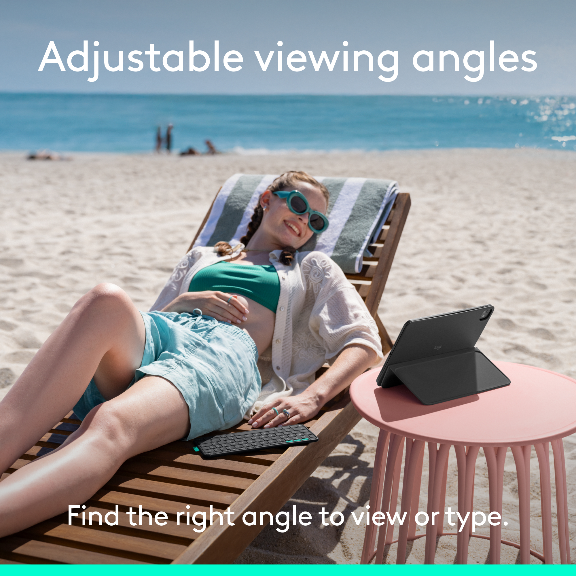 Frau auf Liegestuhl mit Tablet und Tastatur. Strand, Meer und rosafarbener Tisch im Hintergrund.