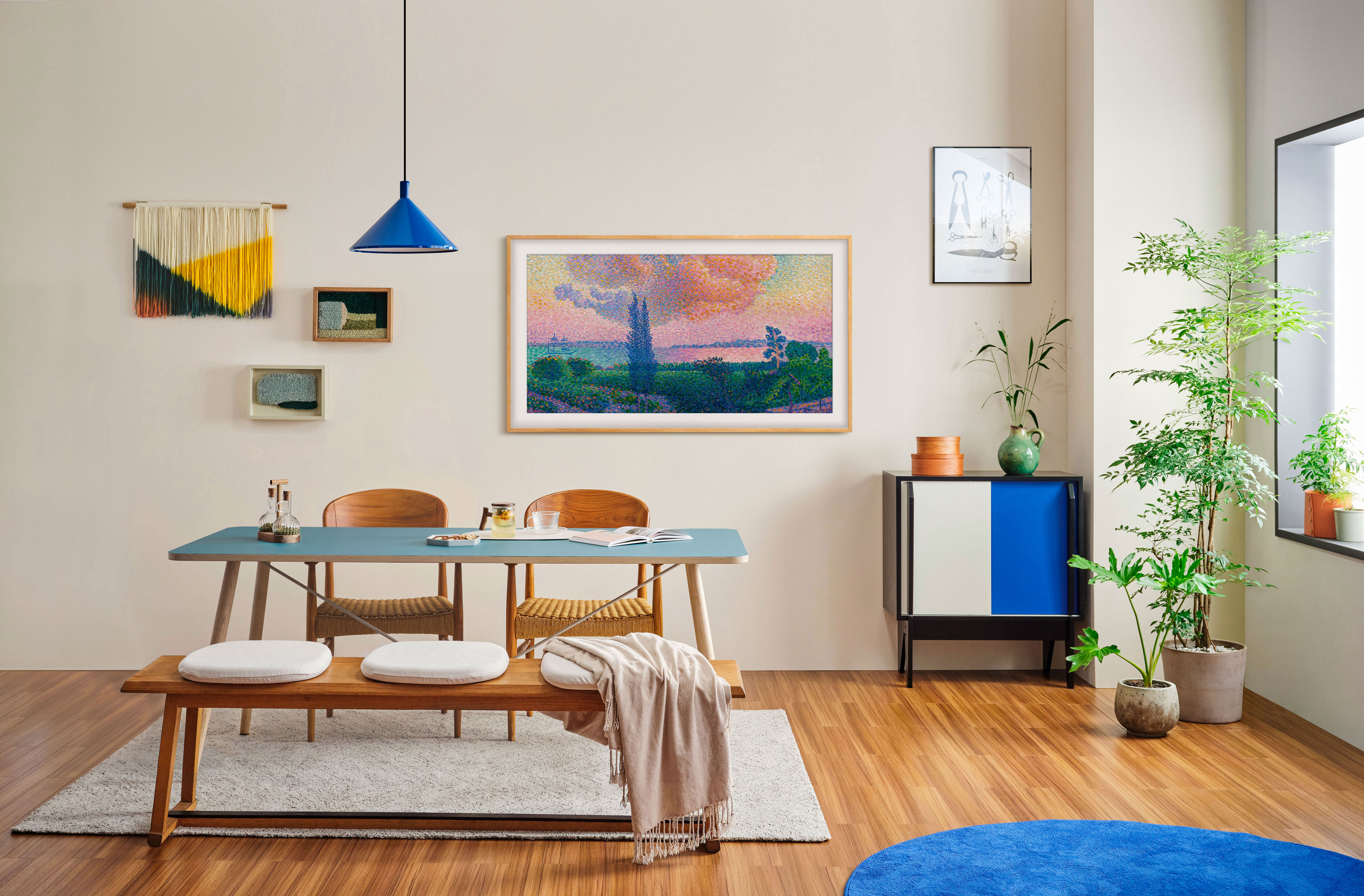 Una sala da pranzo con tavolo blu, opere d'arte, sedie e tappeto blu. Sono visibili anche piante, un mobile e una lampada a sospensione.