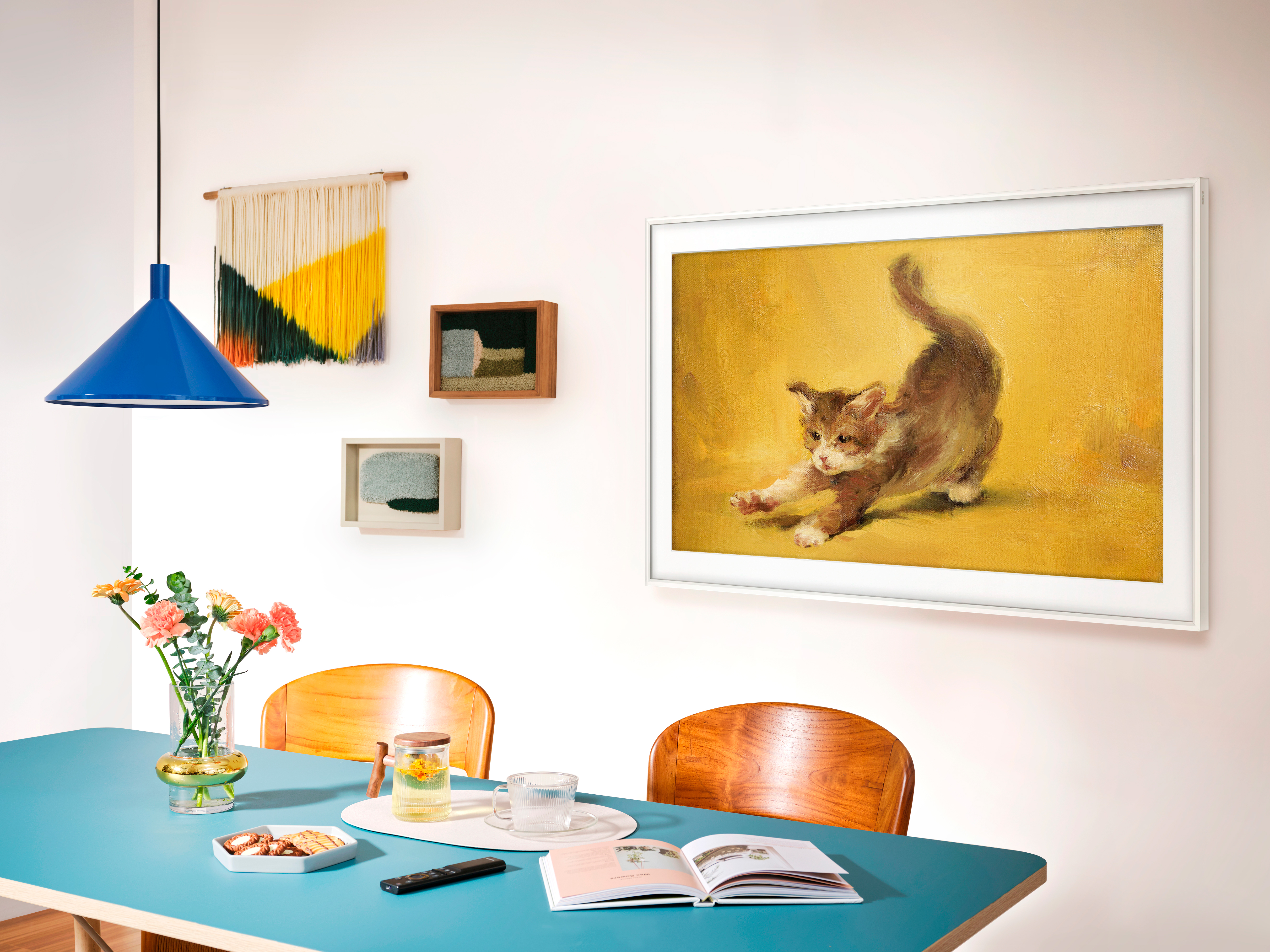 Una stanza con tavolo blu, dipinto di gattino e decorazioni murali. Fiori e lampada visibili.