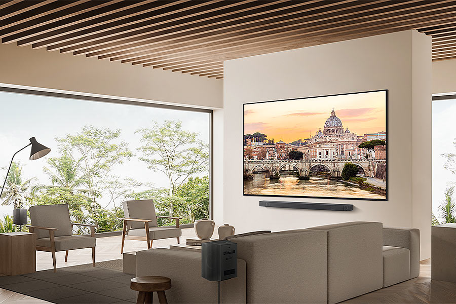 Wohnzimmer mit großem Fernseher, der eine Stadtlandschaft zeigt. Graues Sofa, Stühle und eine schwarze Soundbar.