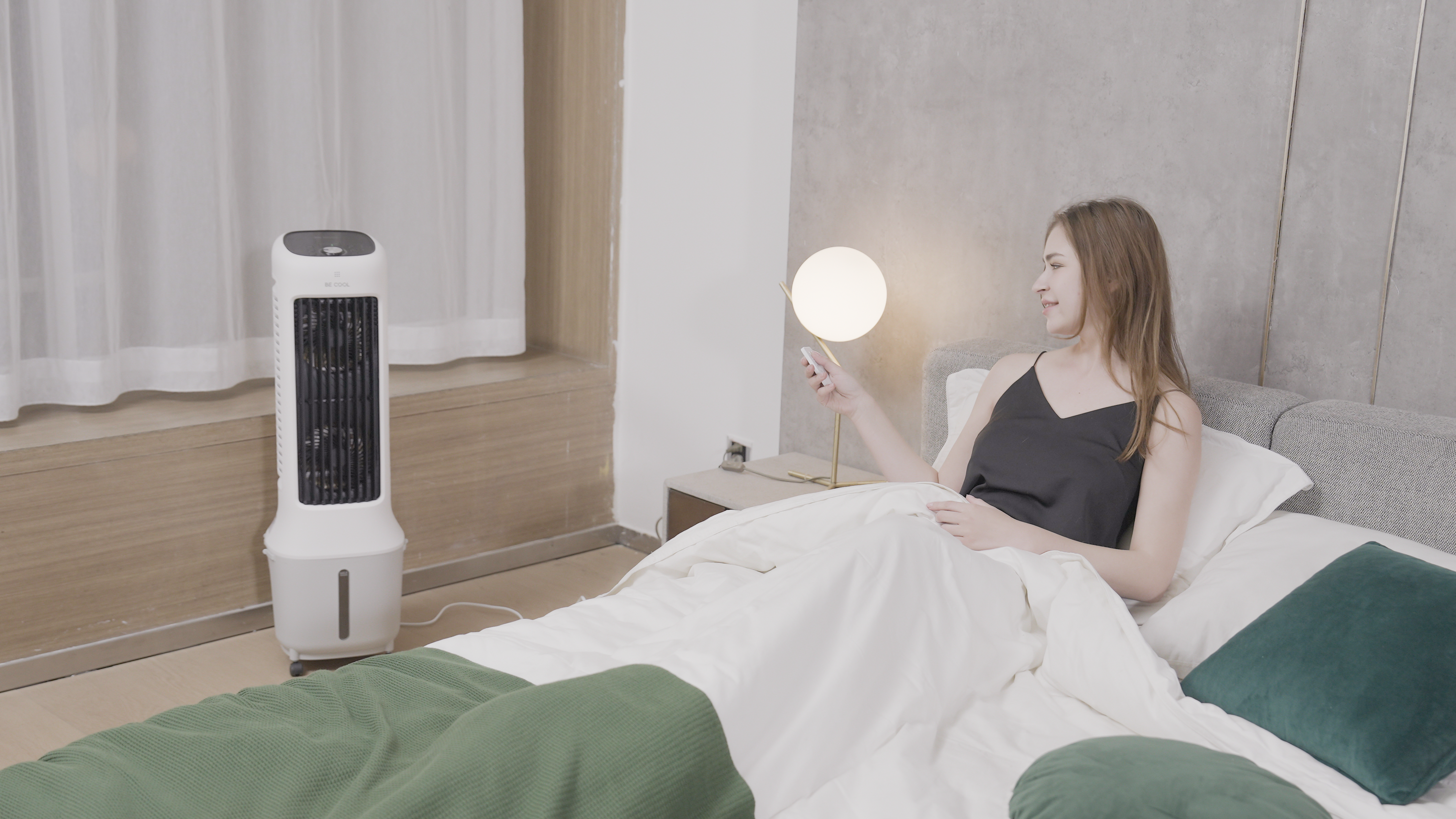 Frau im Bett mit Fernbedienung, neben einer Lampe. Links steht ein weißer Ventilator. Grüne Decke und Kissen sichtbar.