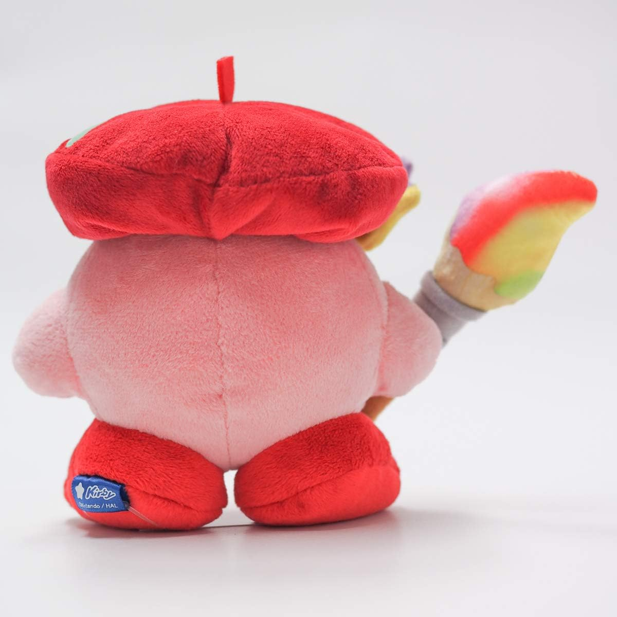 Ein rosa Kirby-Plüschtier mit roter Baskenmütze und buntem Pinsel, rote Füße. Ein Etikett ist angebracht.
