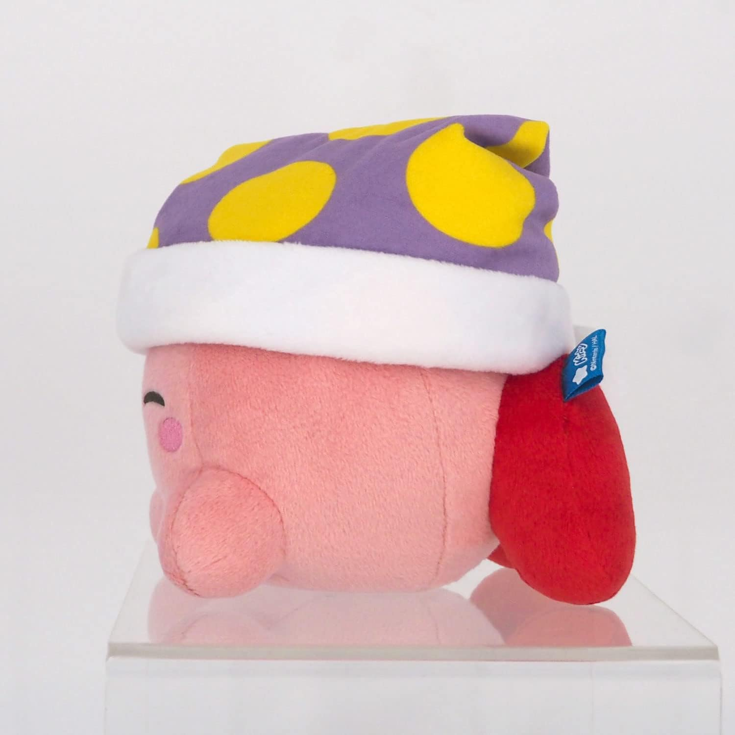 Ein rosa Kirby-Plüschtier mit einem lila Hut mit gelben Punkten und einem weißen Rand. Es steht auf einem klaren Ständer, mit einem roten Anhang.