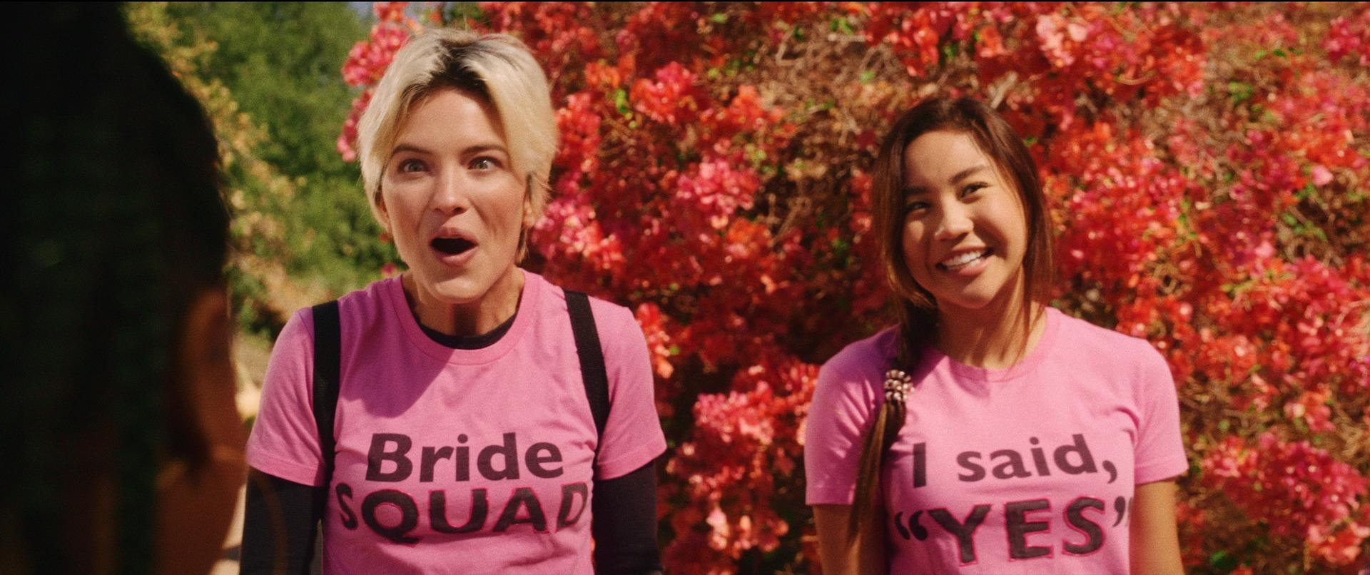 Zwei Frauen in rosa Hemden lächeln und schnappen nach Luft. Eine sagt "Braut Squad", die andere "Ich sagte, JA". Rote Blumen im Hintergrund.