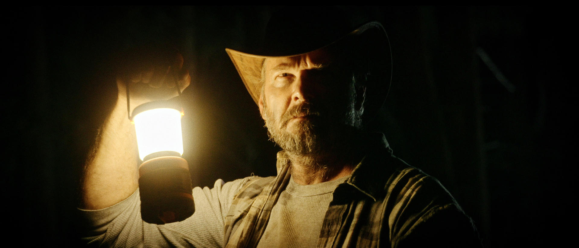 Ein Mann mit Cowboyhut hält eine brennende Laterne in einem dunklen Raum. Sein Gesicht ist beleuchtet und er hat einen Bart. Der Hintergrund ist schwarz.
