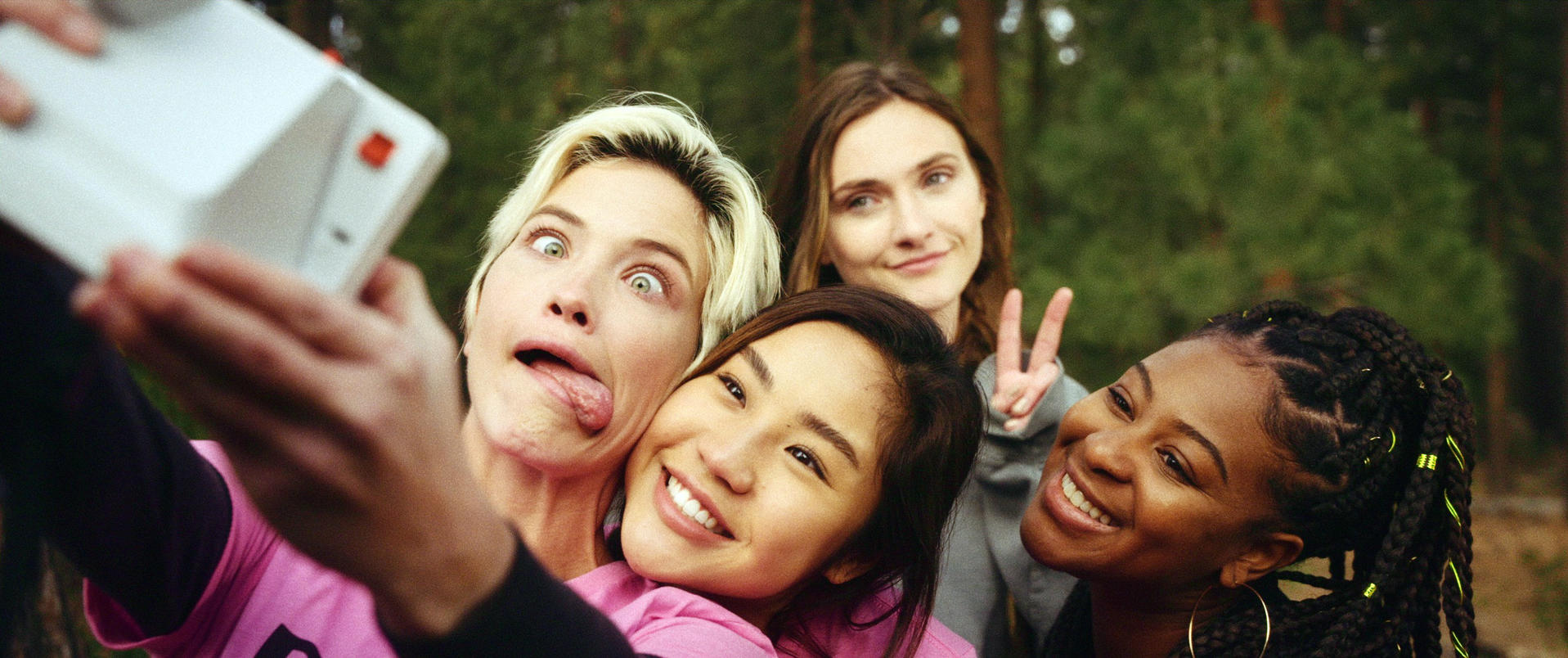 Vier Frauen machen ein Selfie. Eine verzieht das Gesicht, eine andere zeigt ein Friedenszeichen. Bäume im Hintergrund.
