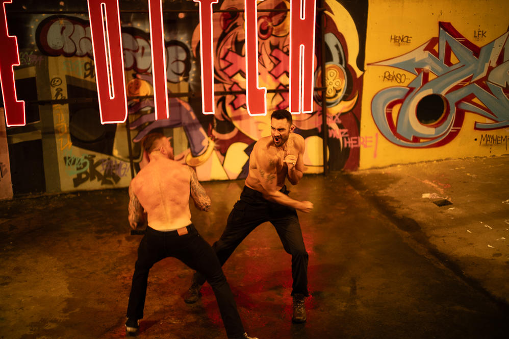 Zwei Männer in Kampfpose, ohne Hemd, vor einer Graffiti-Wand und einem Neon-Schild. Einer trägt schwarze Jeans.