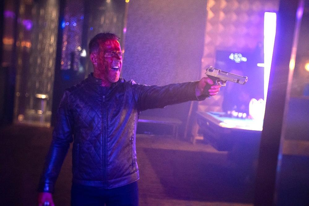 Mann mit Blut im Gesicht, der eine Waffe zielt. Szene ist eine Bar mit Neonlichtern und einem Billardtisch im Hintergrund.