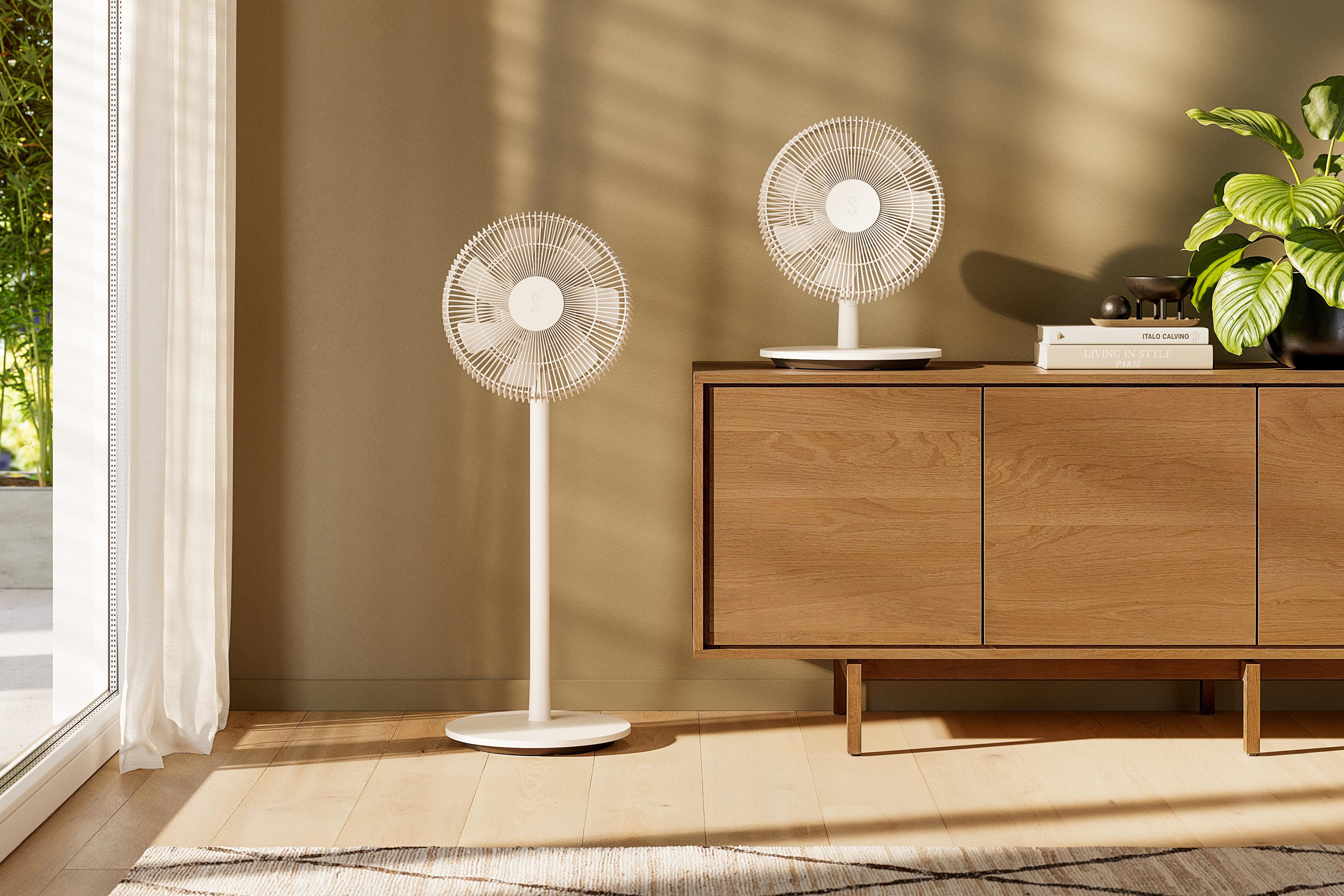Zwei weiße Ventilatoren in einem Raum. Rechts ein Holzschrank. Licht scheint durch das Fenster.