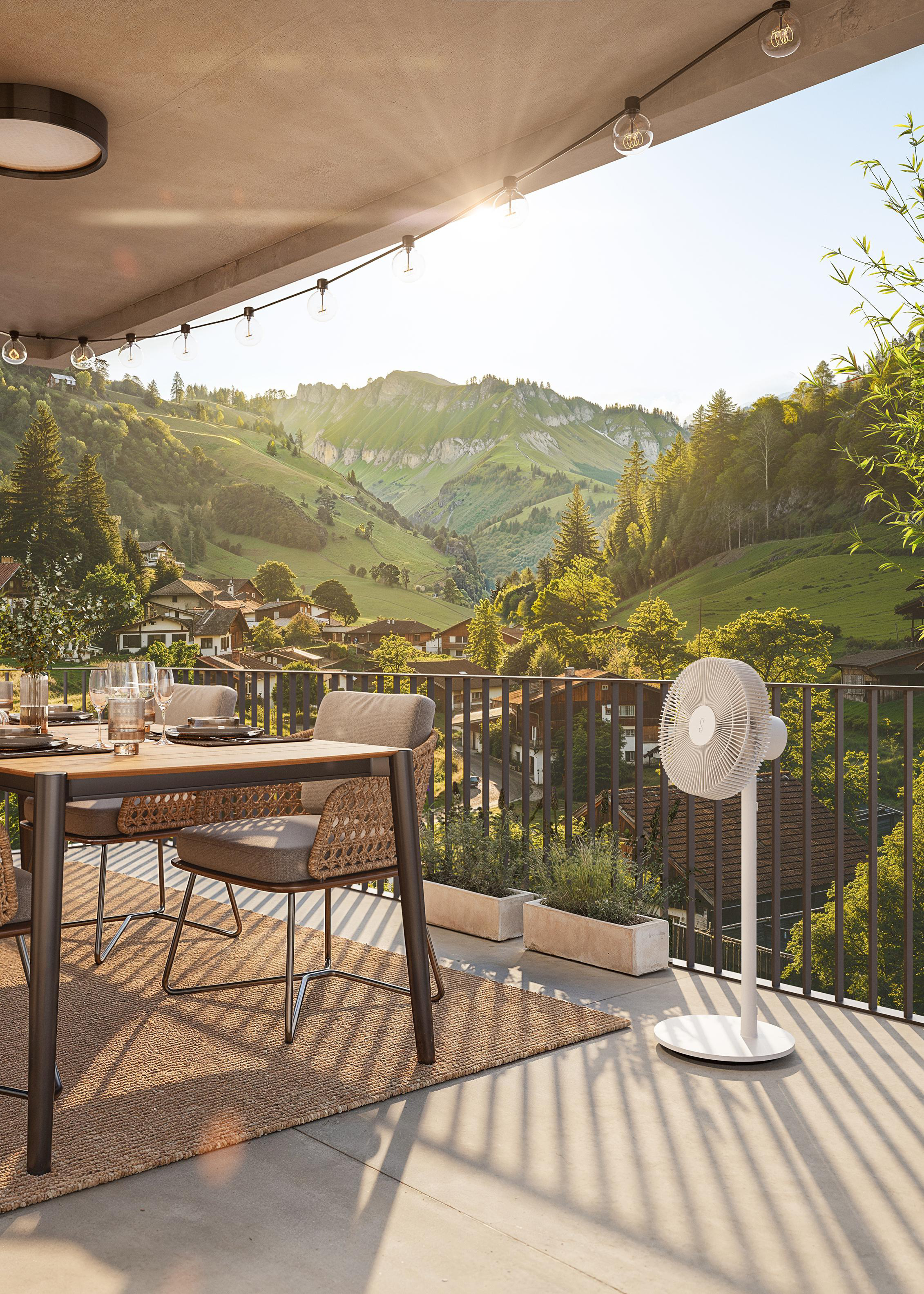 Balkon mit Tisch, Essgeschirr, Ventilator und Blick auf grüne Hügel und Häuser. Lichterketten hängen oben.
