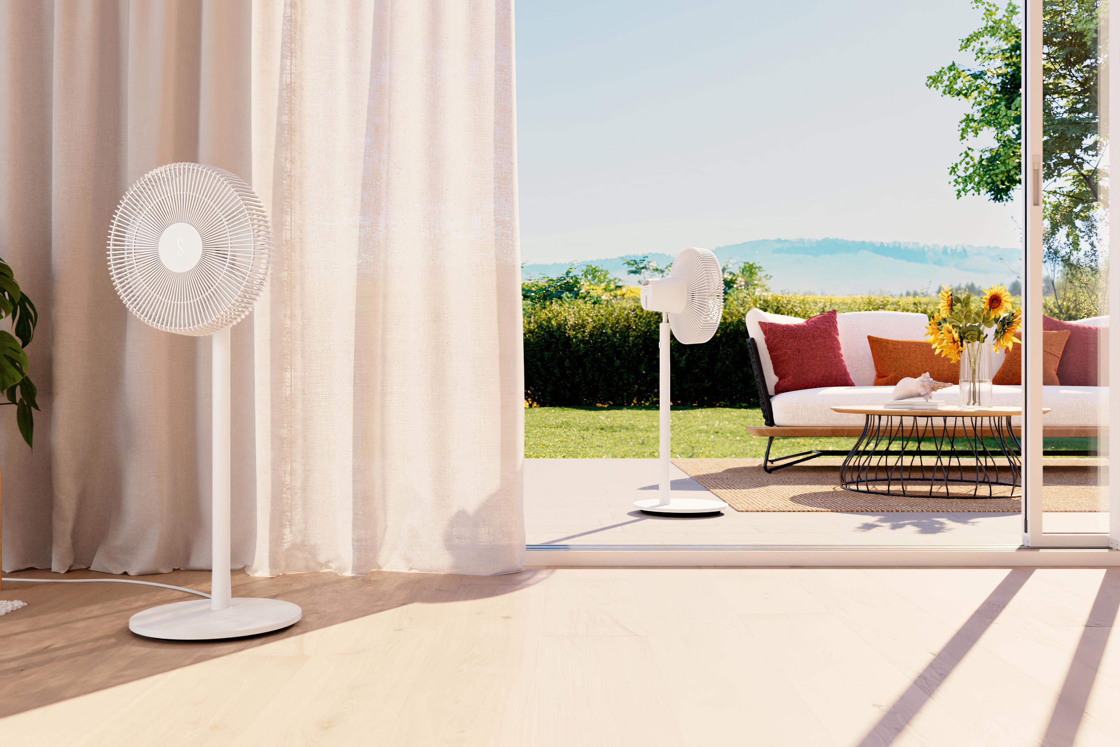 Weiße Ventilatoren und Vorhänge, offene Tür zu Terrasse mit Sofa, Tisch und Blumen. Blauer Himmel und grüne Landschaft.