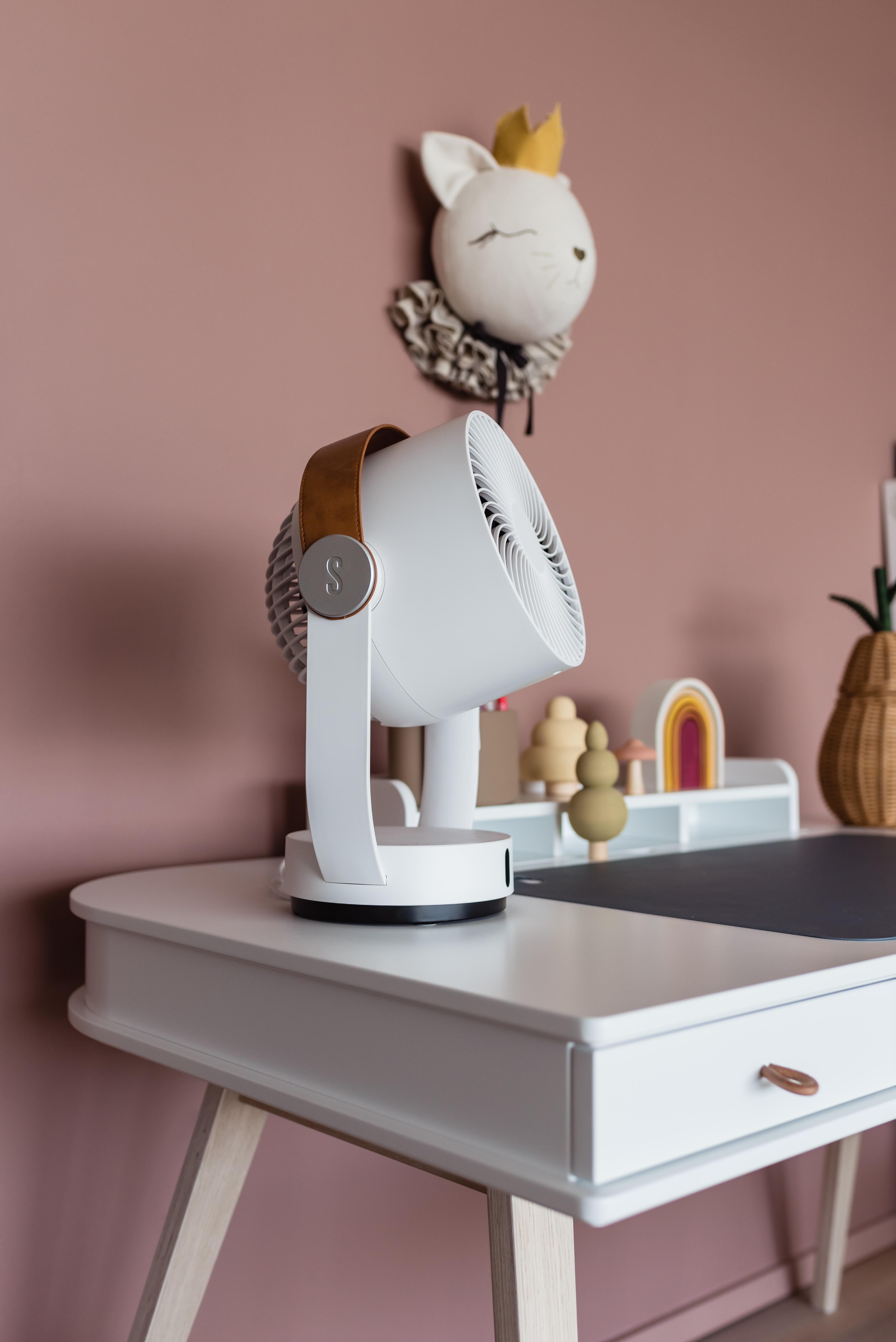 Weißer Schreibtischventilator mit braunem Griff steht auf einem weißen Schreibtisch. Eine Katzenkopf-Deko ziert die rosa Wand.
