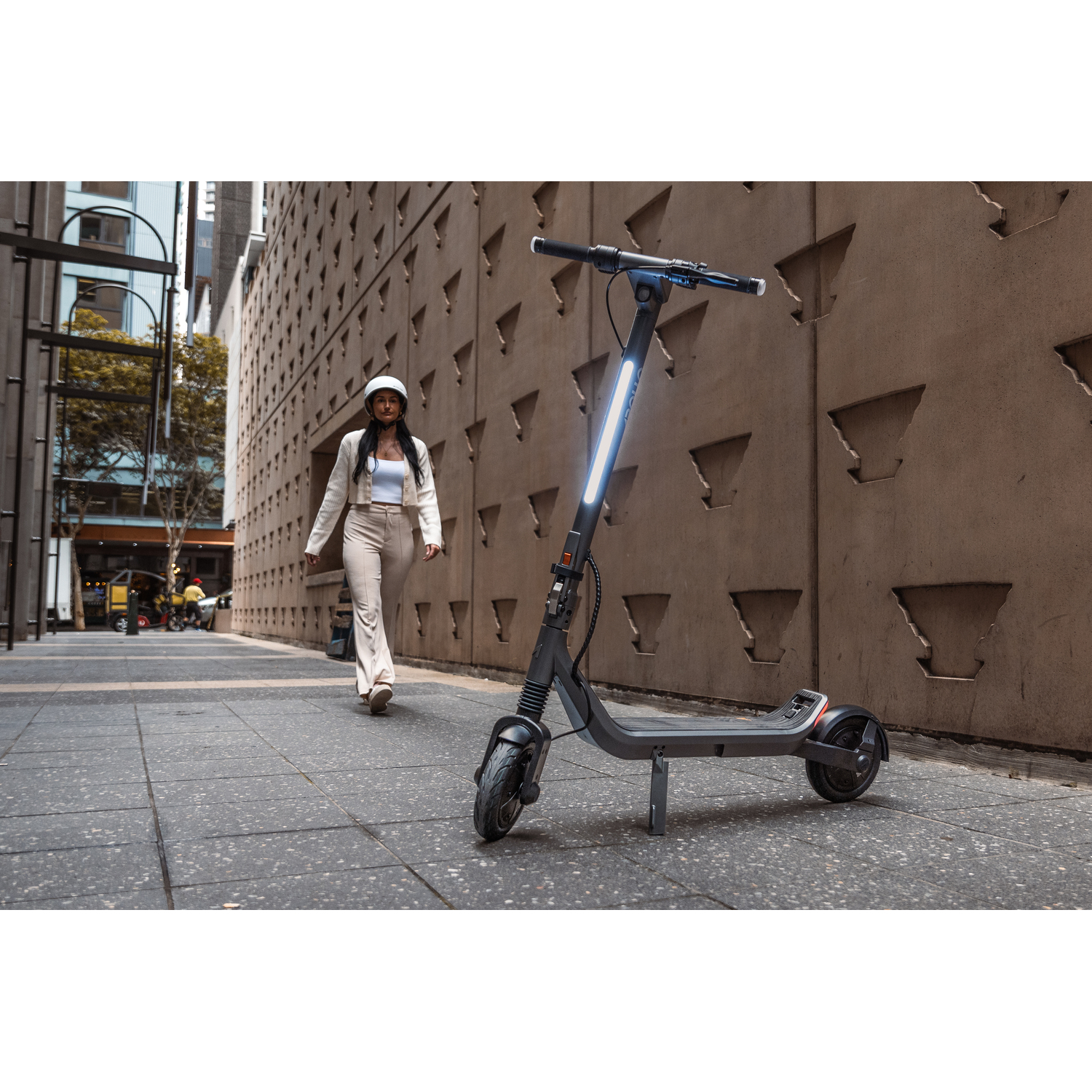 Une femme en beige passe devant une trottinette électrique. La trottinette est sur le trottoir. Derrière, un mur en béton texturé.