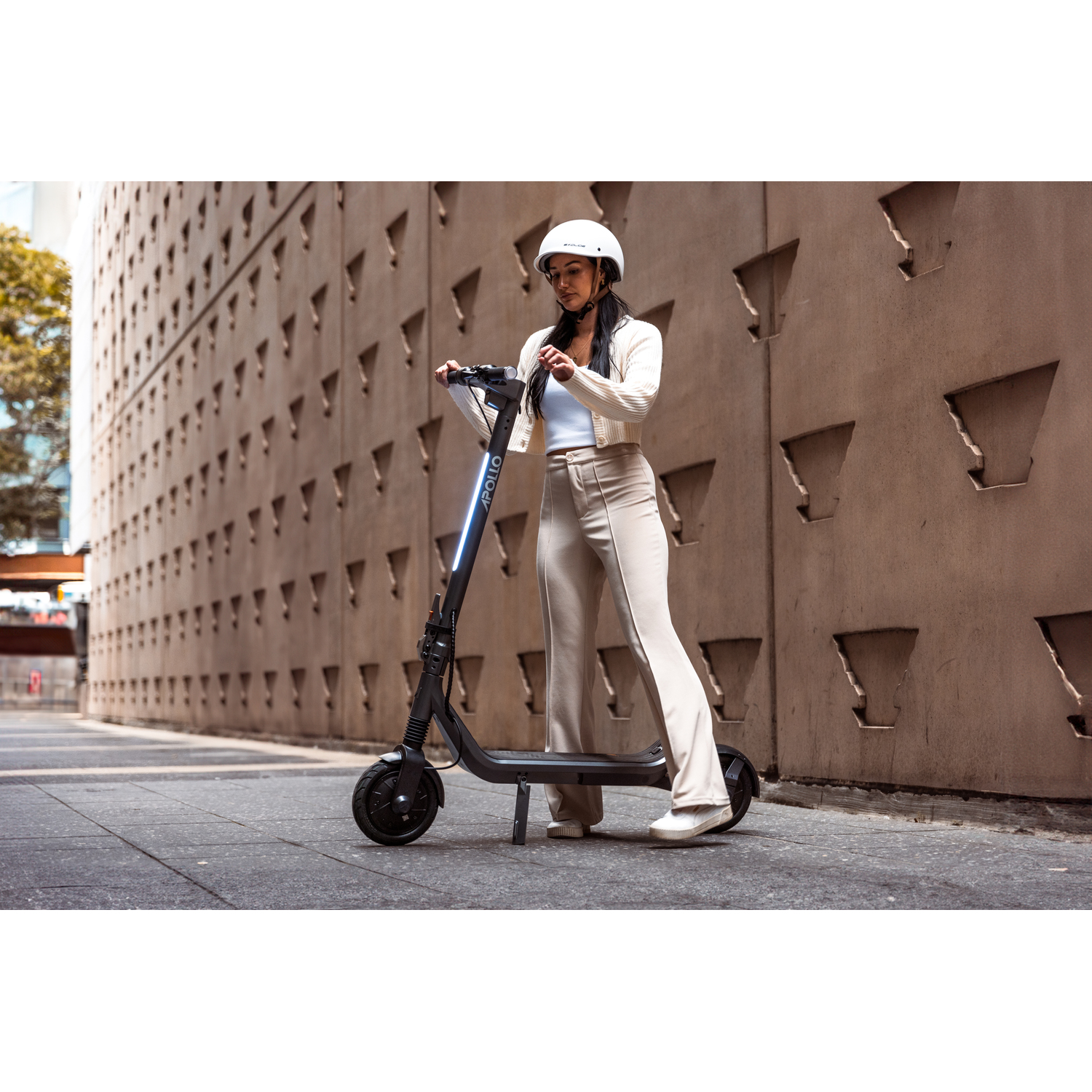 Une femme portant un casque blanc se tient debout avec une trottinette Apollo noire. Elle porte un pantalon et une veste beiges. Un bâtiment texturé en arrière-plan.