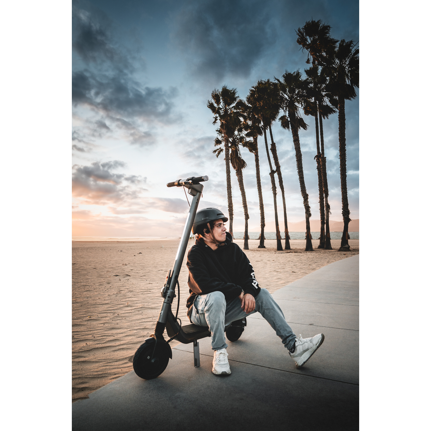Une personne assise sur un scooter près de la plage, portant un casque. Palmiers et ciel nuageux en arrière-plan. Teintes de coucher de soleil.