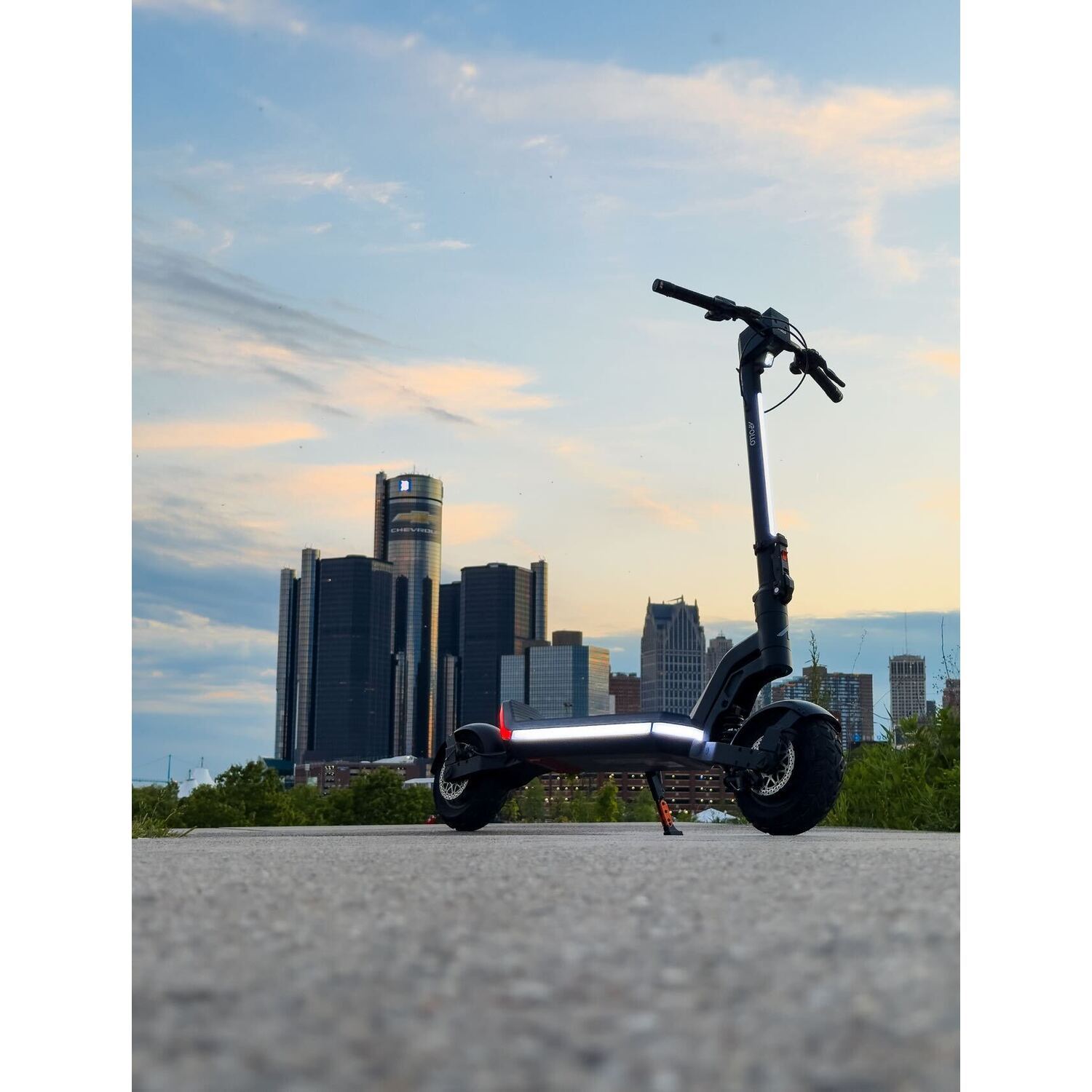 An electric scooter stands on a concrete path with a city skyline in the background at dusk. The sky is blue and orange.