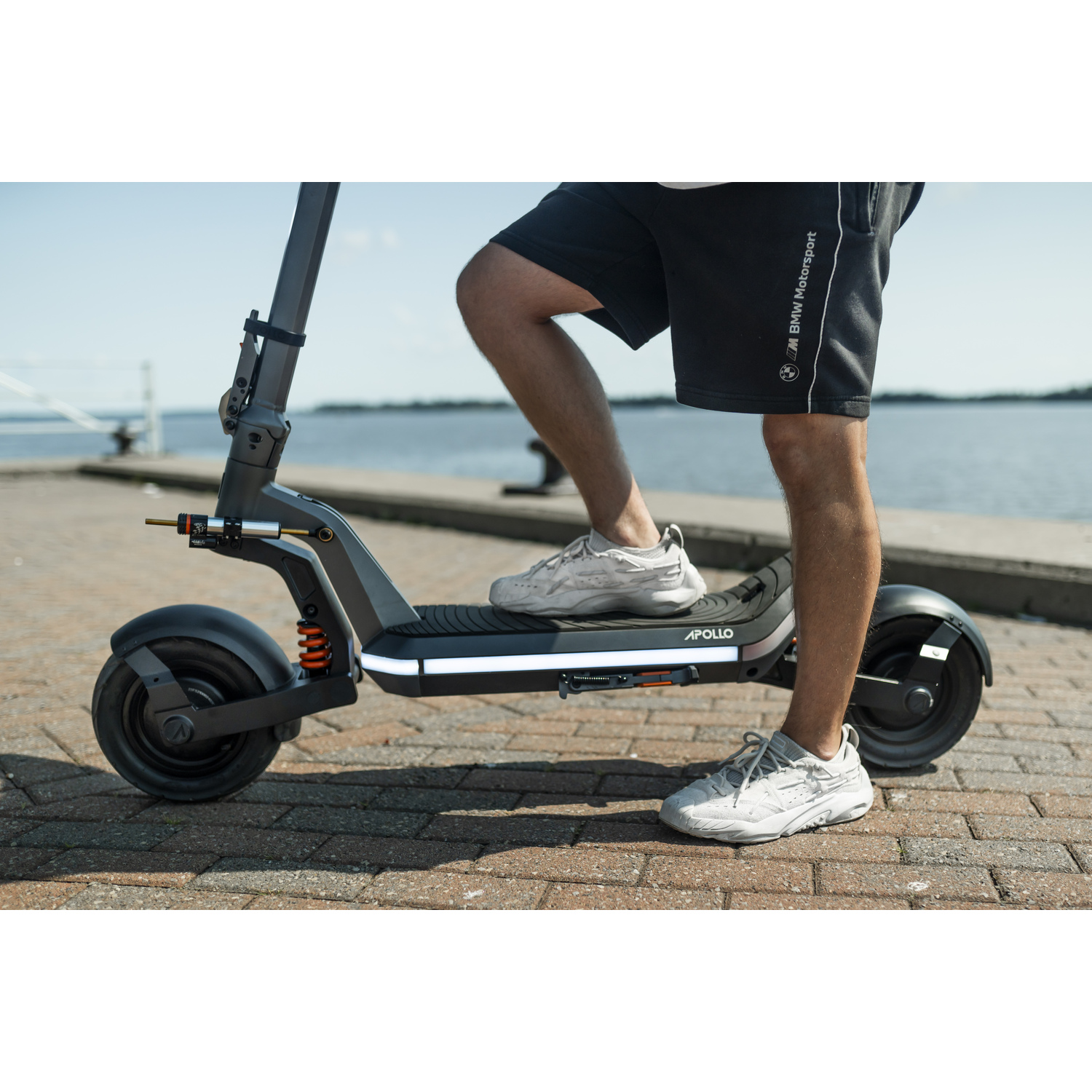 Person on an Apollo scooter, legs visible. Black shorts, grey shoes. Background shows water, a bridge, and a brick walkway.