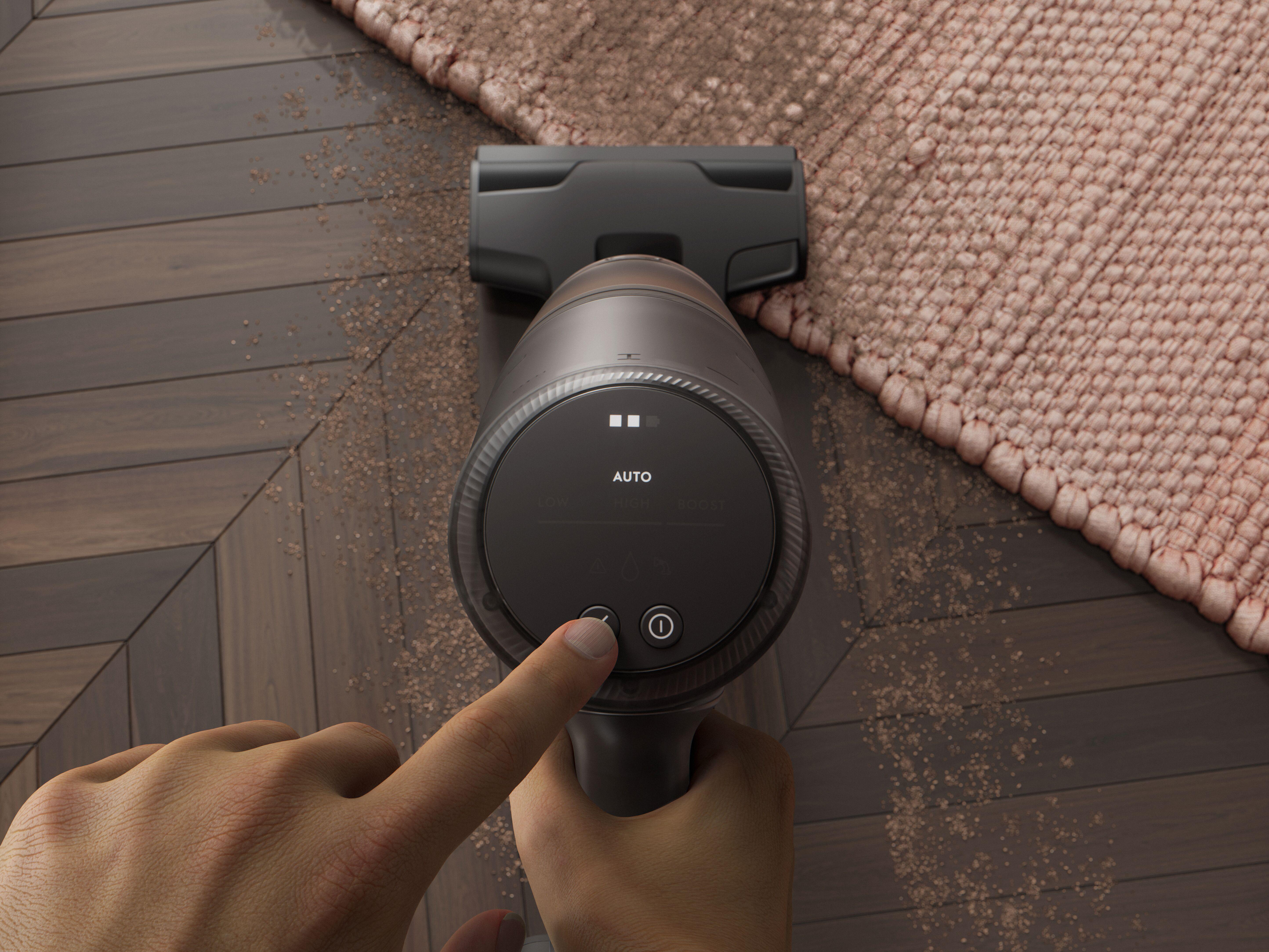 Eine Hand drückt eine Taste an einem schwarzen Staubsauger. Der Staubsauger steht auf einem Holzboden mit einem Teppich. Staub ist sichtbar.