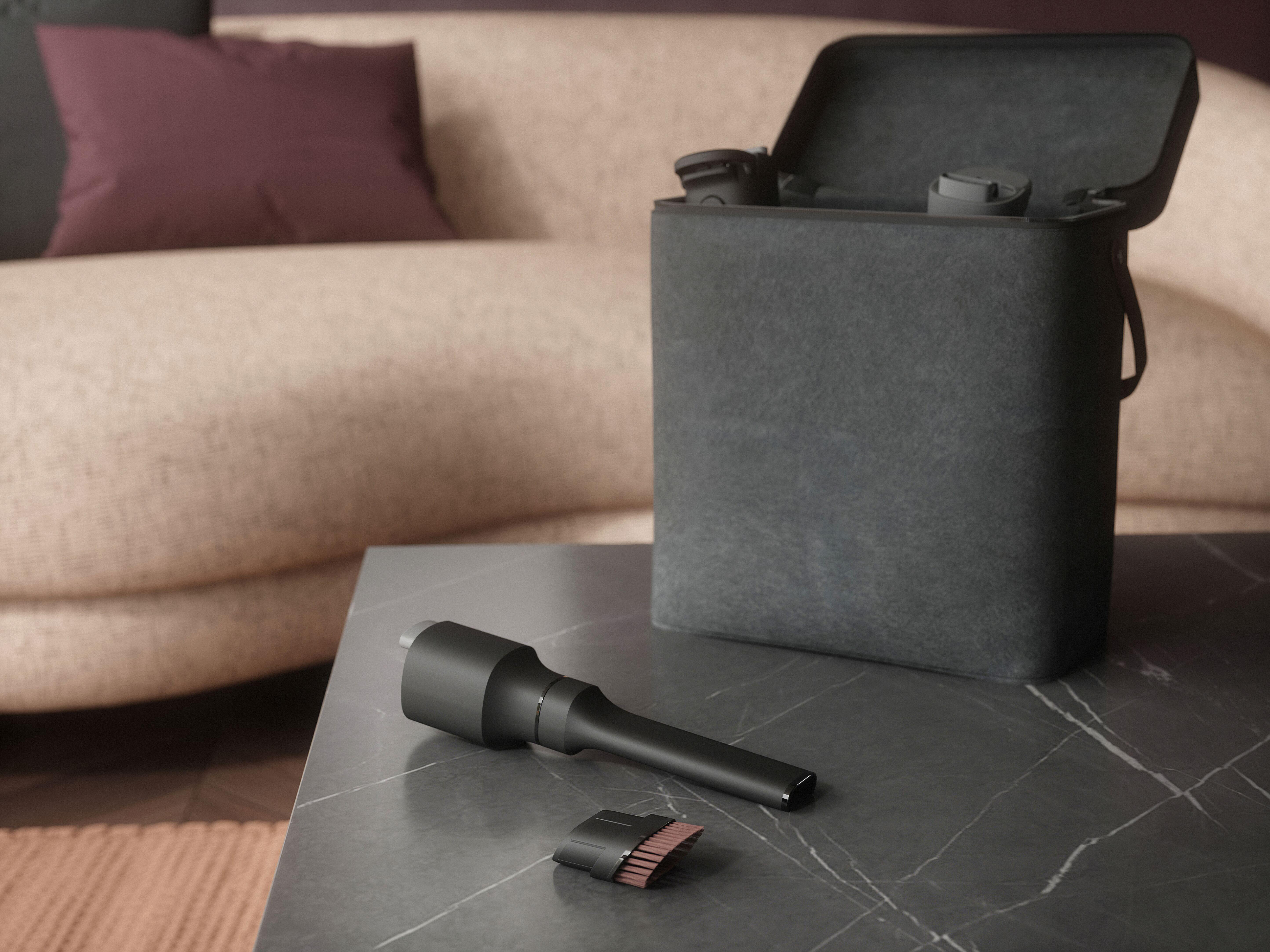 Ein schwarzer Staubsauger und Zubehör liegen auf einem Marmortisch, im Hintergrund eine Tasche neben einem Sofa.