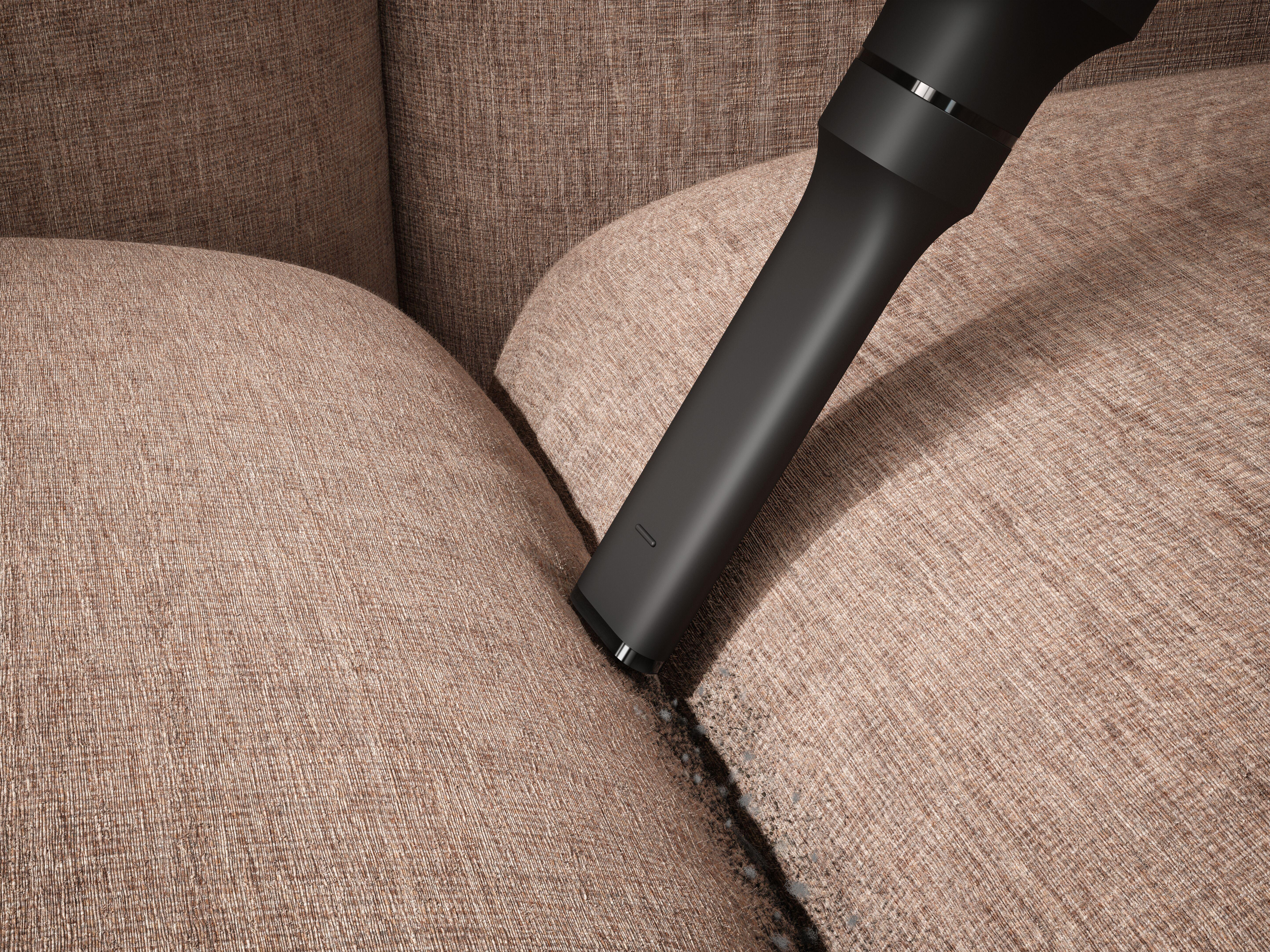 Eine schwarze Staubsaugerdüse reinigt ein beigefarbenes Stoffsofa. Das Sofa hat eine sichtbare Textur und wird von oben beleuchtet.
