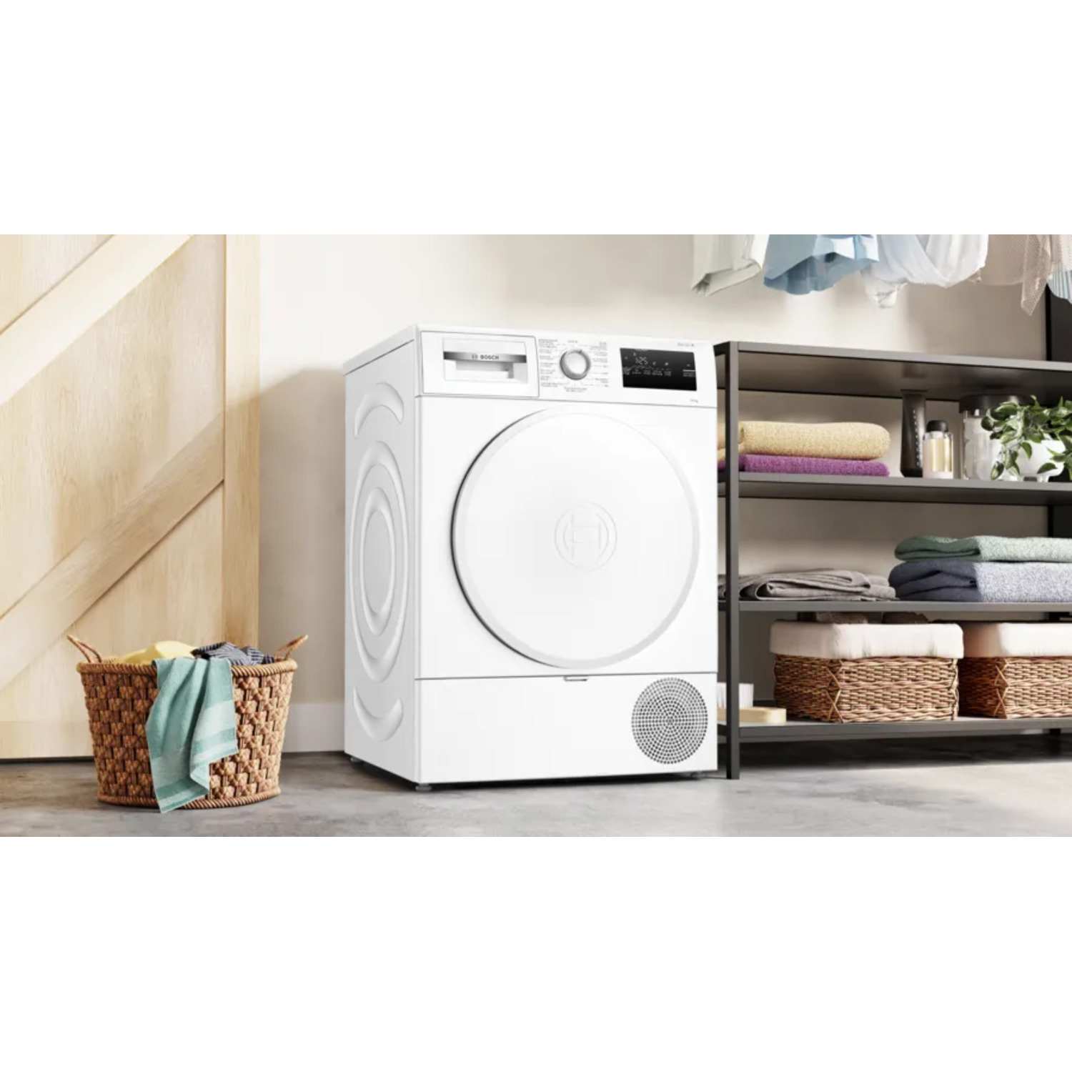 Un sèche-linge blanc est dans une buanderie à côté d'un panier de linge. Des étagères contiennent des serviettes et des paniers. Porte en bois à gauche.