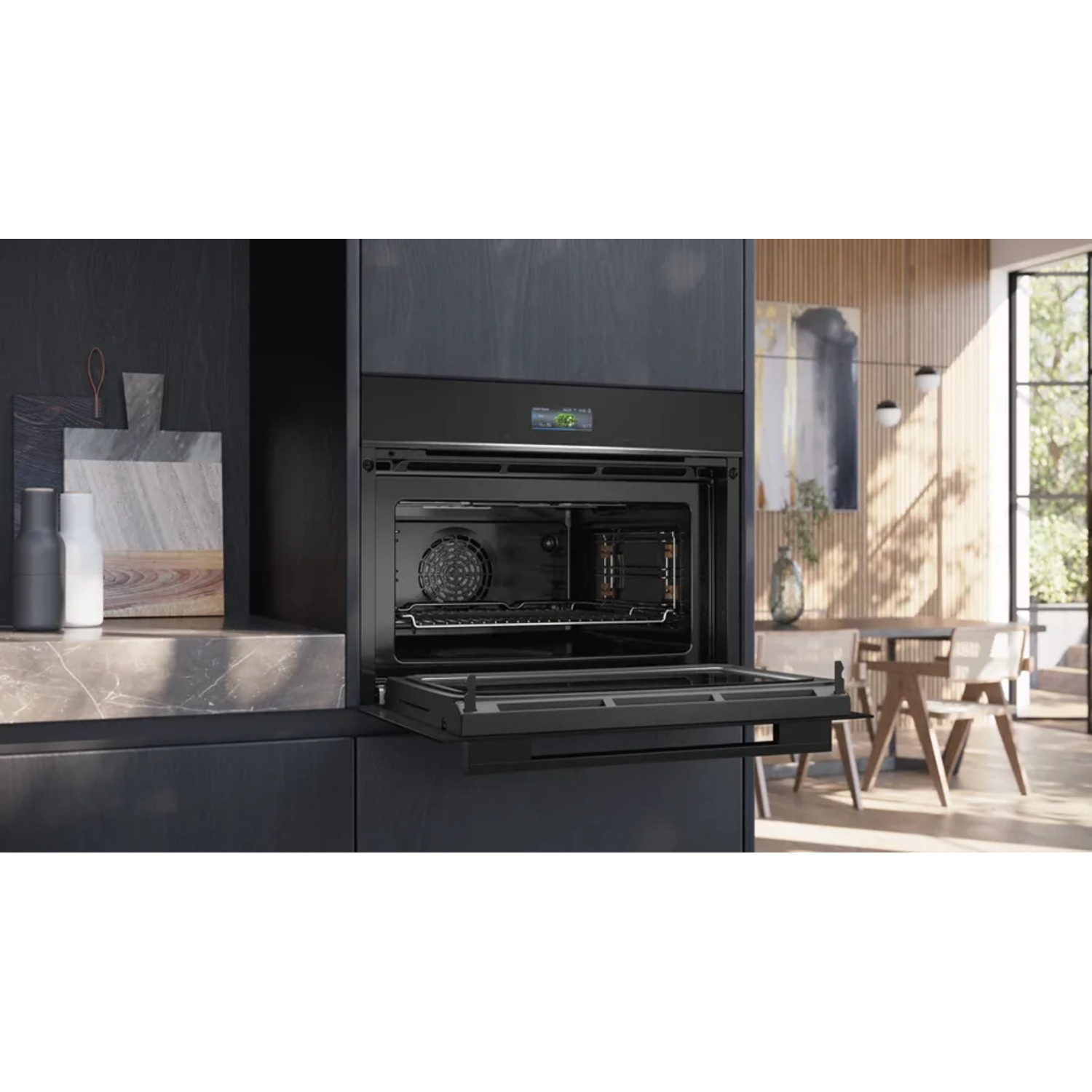 Black oven door open, revealing interior with fan and racks. Kitchen setting with cutting boards and a dining area visible.