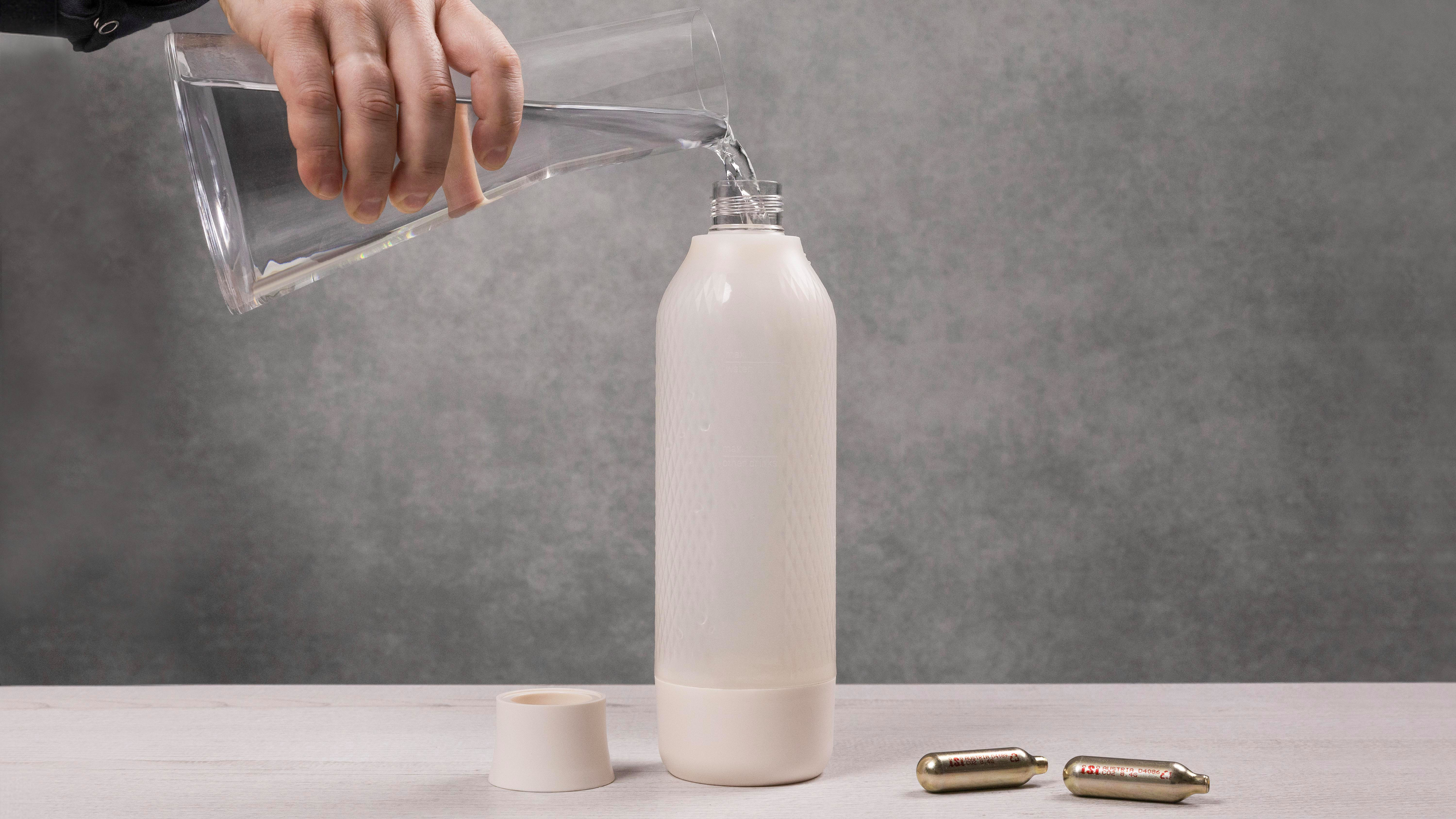 Eine Hand gießt Wasser aus einem Glas in eine weiße Flasche. Eine Kappe und zwei kleine Kanister liegen auf dem Tisch.