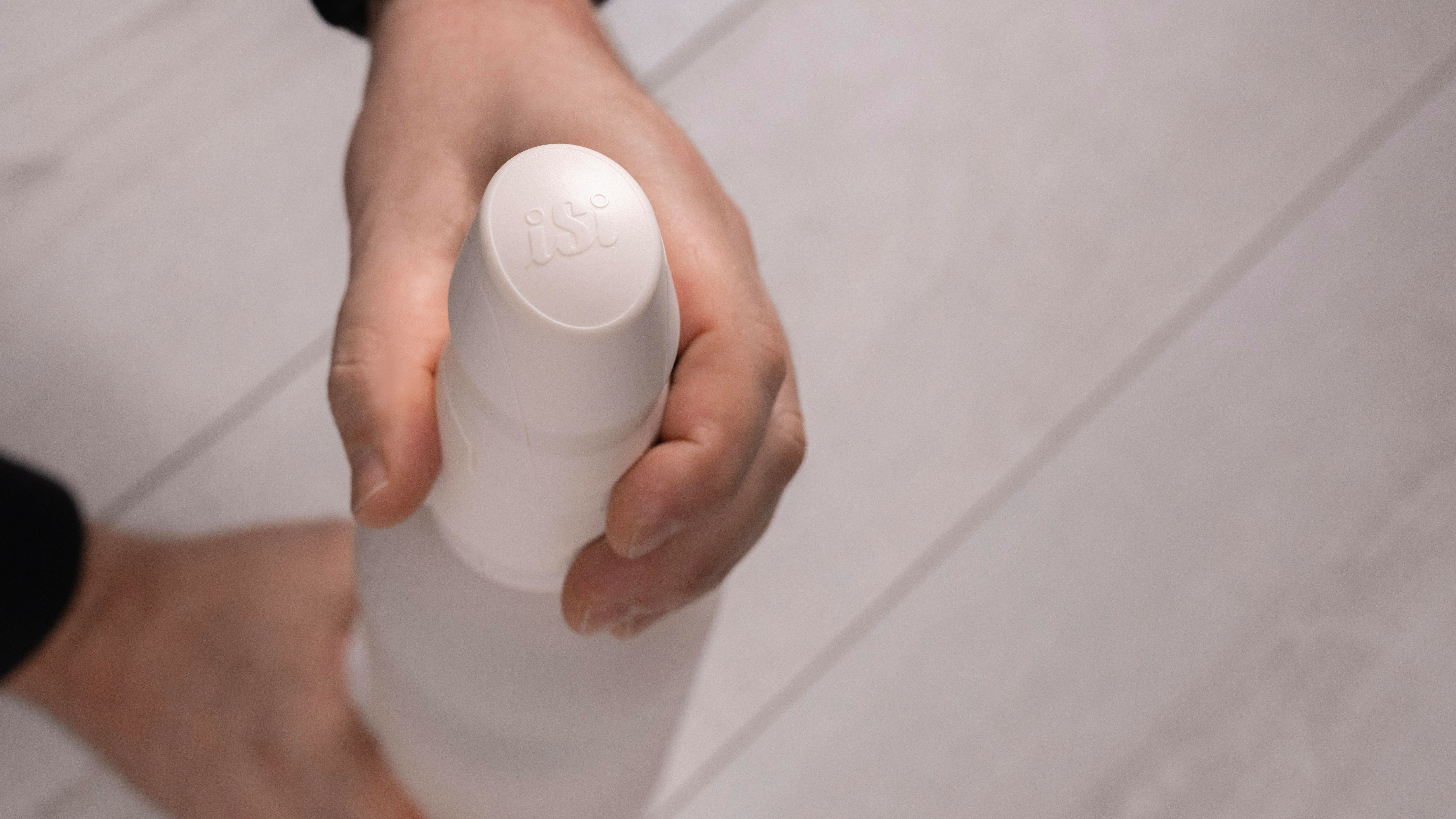 Eine Hand hält eine weiße Sprühflasche mit den Buchstaben „ist“ oben. Der Hintergrund ist eine helle Holzoberfläche.
