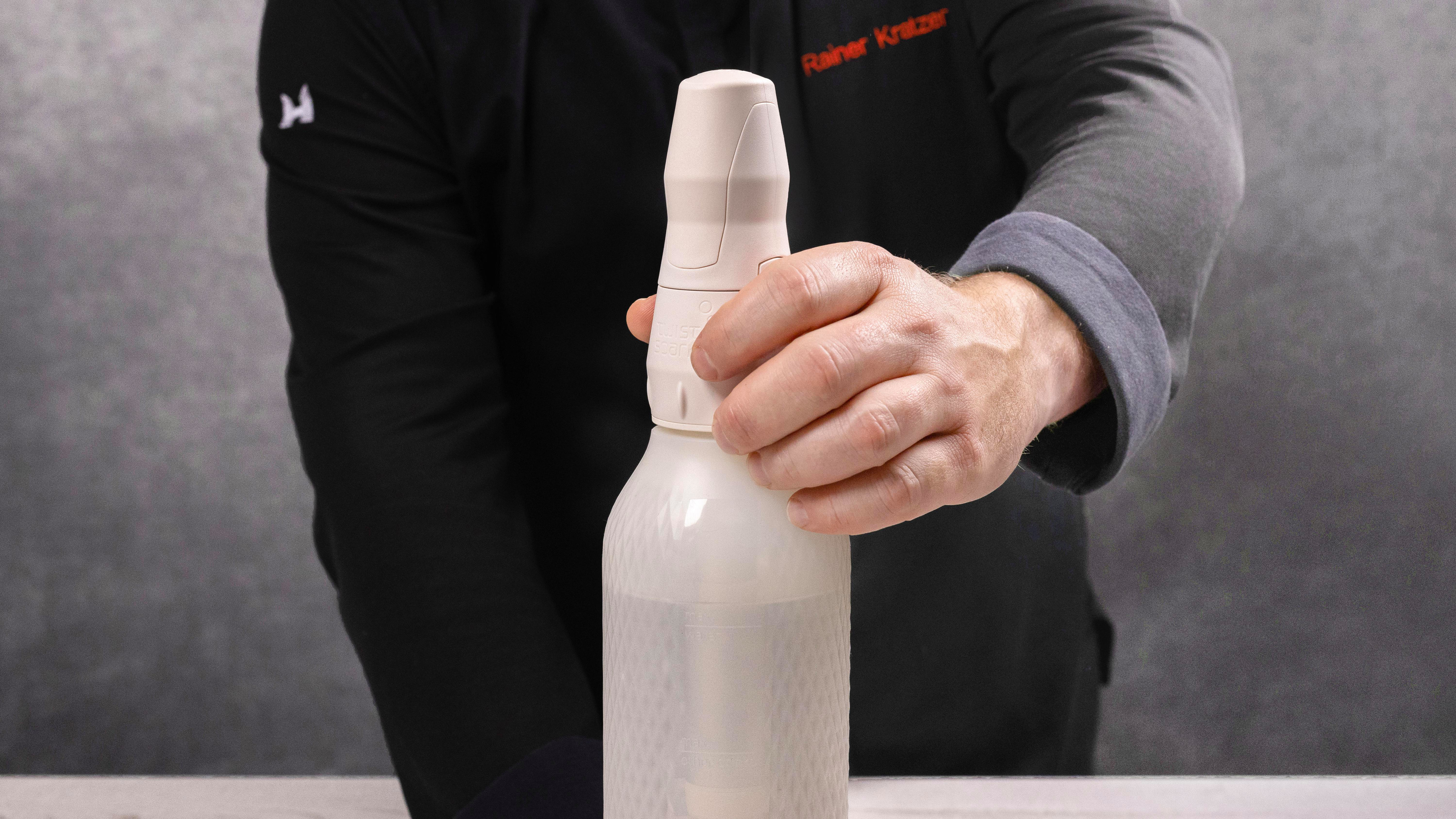 Eine Person hält eine weiße Sprühflasche. Die Person trägt ein schwarzes Hemd und einen grauen Ärmel. Der Hintergrund ist eine graue Wand.