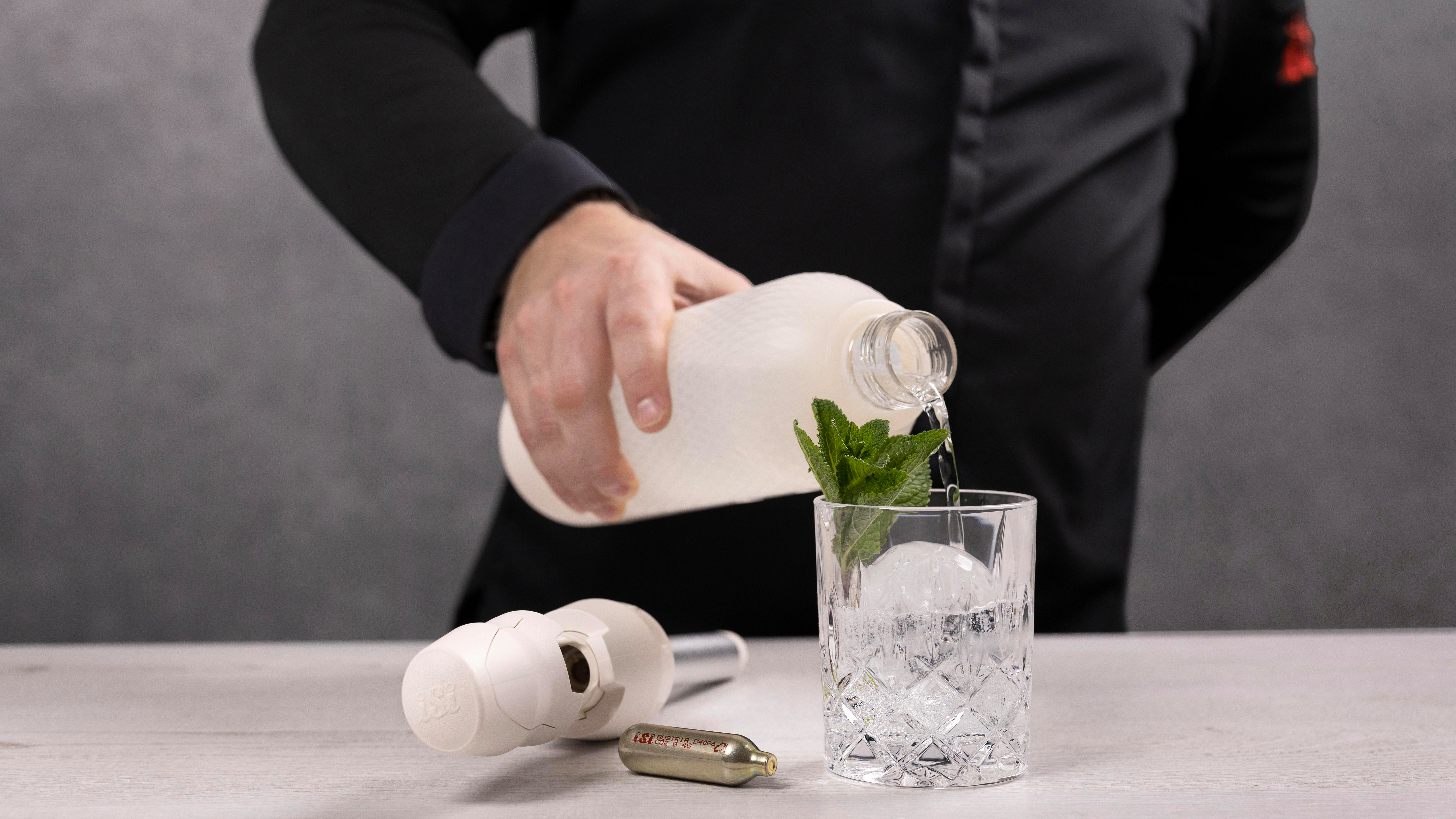 Eine Person gießt Flüssigkeit aus einer weißen Flasche in ein Glas mit Eis und Minze. Bartending-Werkzeuge liegen auf dem Tisch.