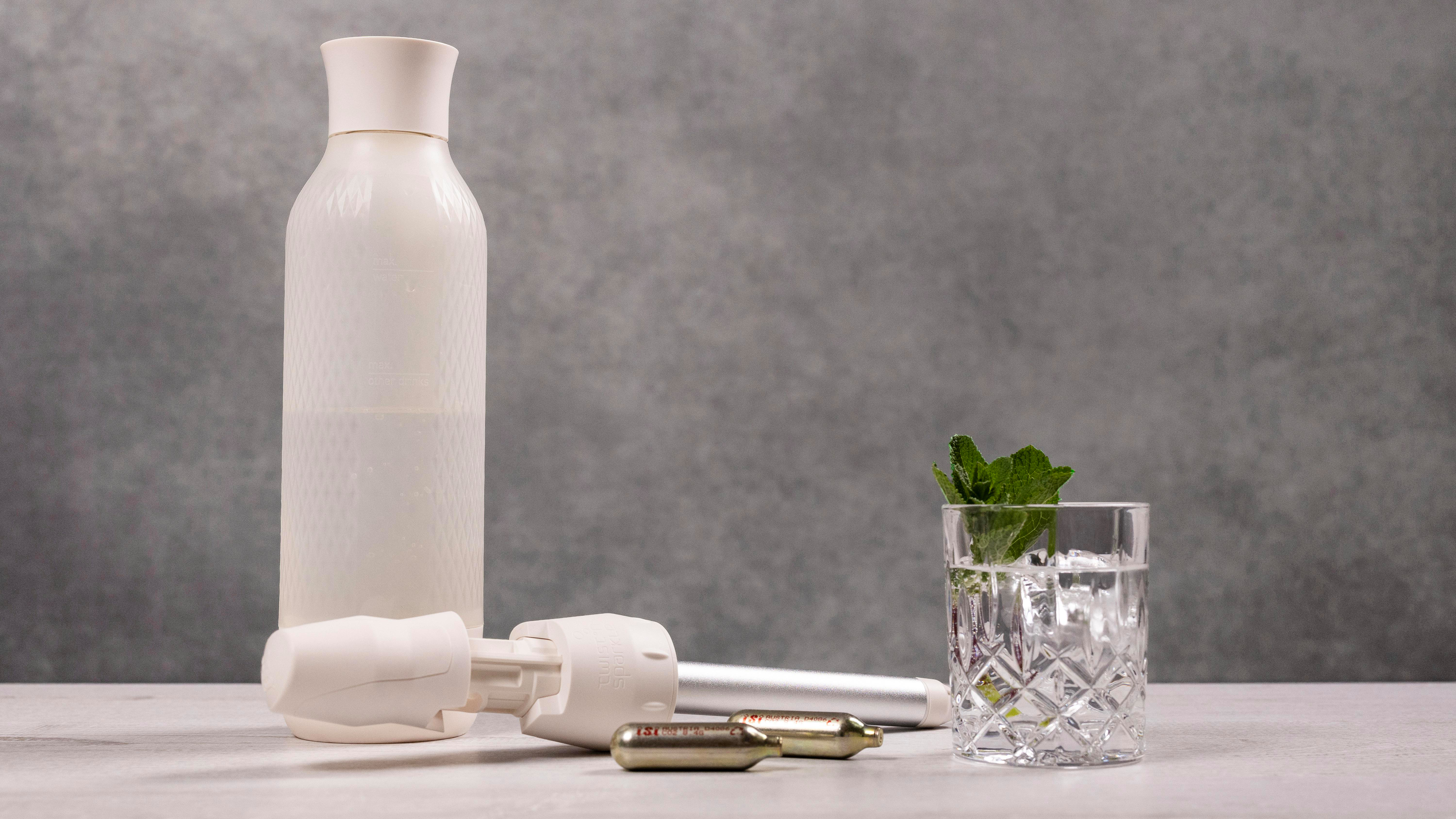 Eine weiße Wasserflasche, ein Sprudelbereiter, ein Glas Wasser mit Eis und Minze sowie CO2-Kartuschen liegen auf einer Oberfläche.