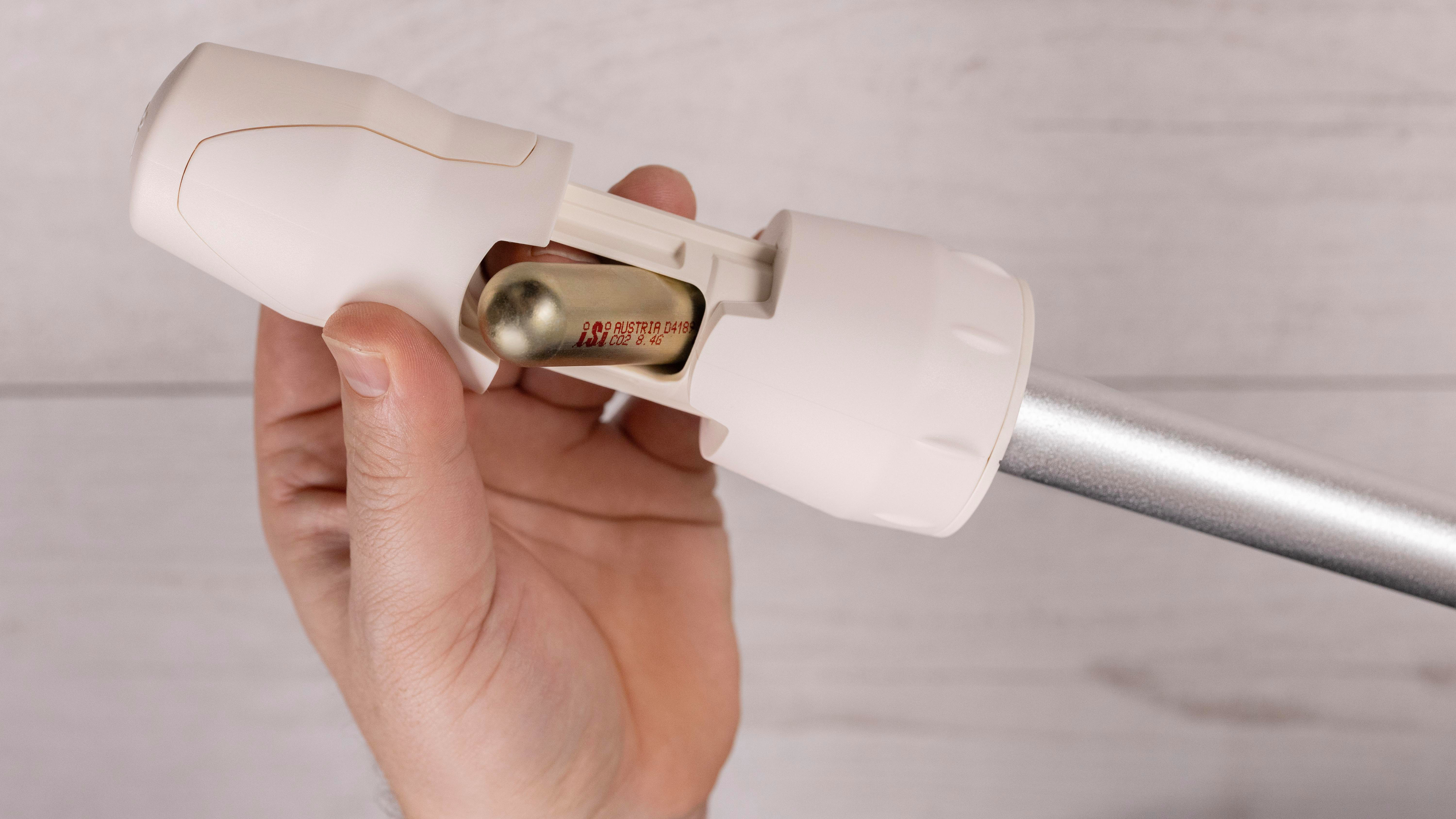 Eine Hand hält ein weißes Gerät mit einem silbernen Rohr. Eine goldene CO2-Patrone ist sichtbar. Der Hintergrund ist eine helle Holzoberfläche.