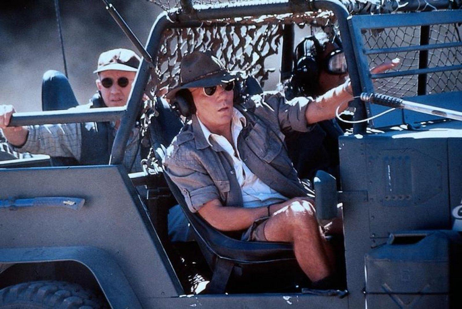 Männer in einem Jeep. Einer trägt Hut und Sonnenbrille. Ein anderer trägt eine Mütze. Sie sitzen in einem Fahrzeug mit Käfig und Tarnnetz.