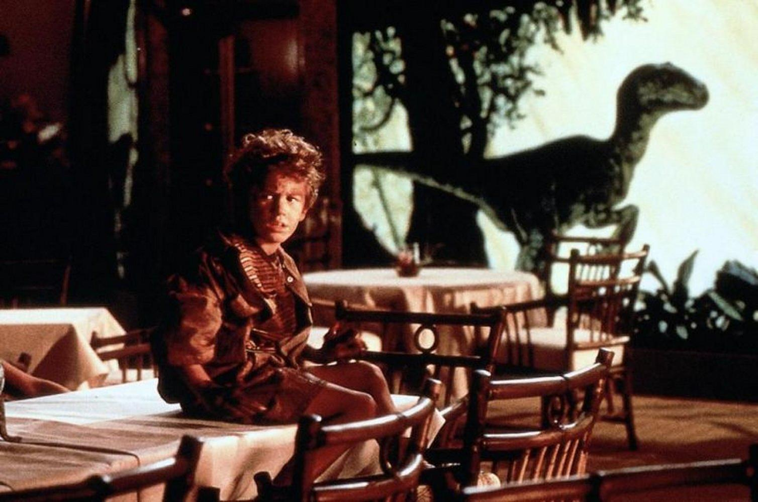 Ein Junge sitzt in einem Restaurant auf einem Tisch. Im Hintergrund eine Dinosaurier-Silhouette. Tische und Stühle füllen die Szene.
