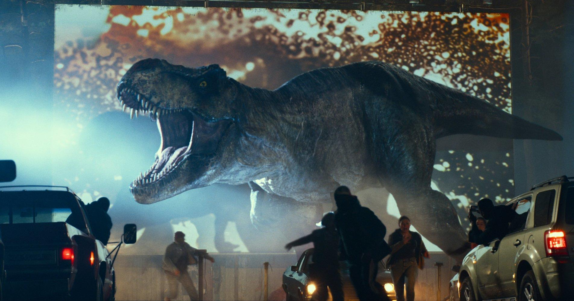 Ein großer T-Rex brüllt vor einer Leinwand. Menschen rennen vor Autos im Vordergrund. Die Szene wirkt chaotisch und gefährlich.